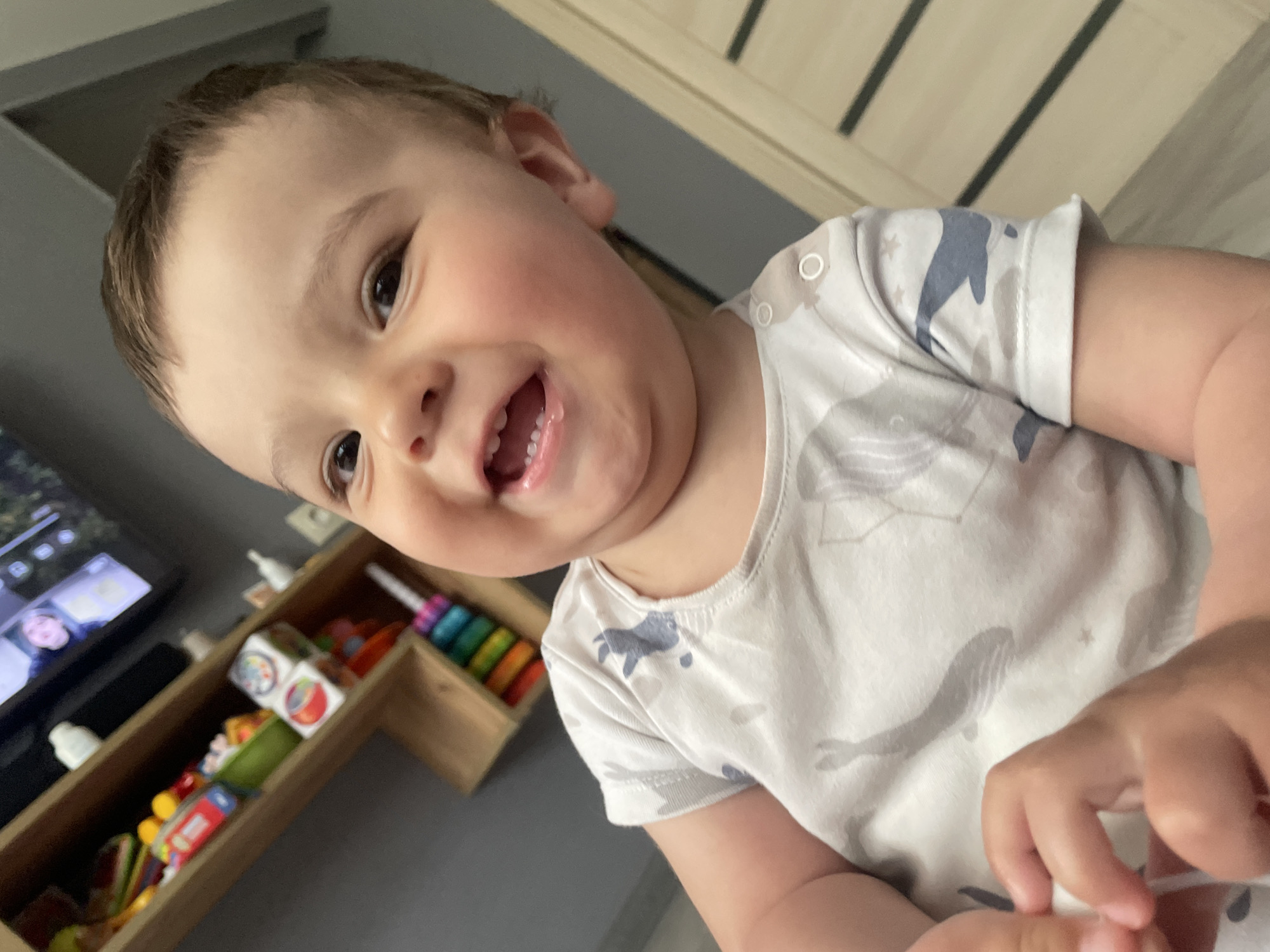 A baby is looking at the camera and smiling. The baby is wearing a light-colored short-sleeved shirt with a whale and star pattern. In the background, there is a television, a wooden organizer filled with toys, and a door with a vertical panel design. No location in Chaikovsky, Russia can be determined from the image.