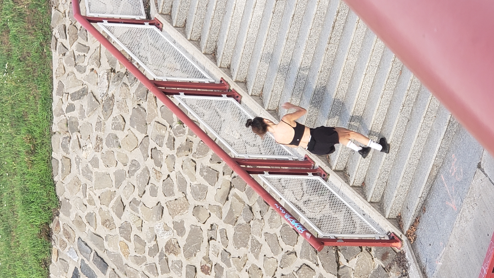 The image shows a woman with dark hair in a bun, wearing a black sports bra and black shorts, with white socks and black athletic shoes, running up a concrete staircase. The staircase has red handrails and white metal mesh railings. Behind the staircase is a stone wall topped with green grass. The scene appears to be outdoors, possibly in a park or recreational area. There are no other people or animals visible in the image. The lighting suggests it is daytime.