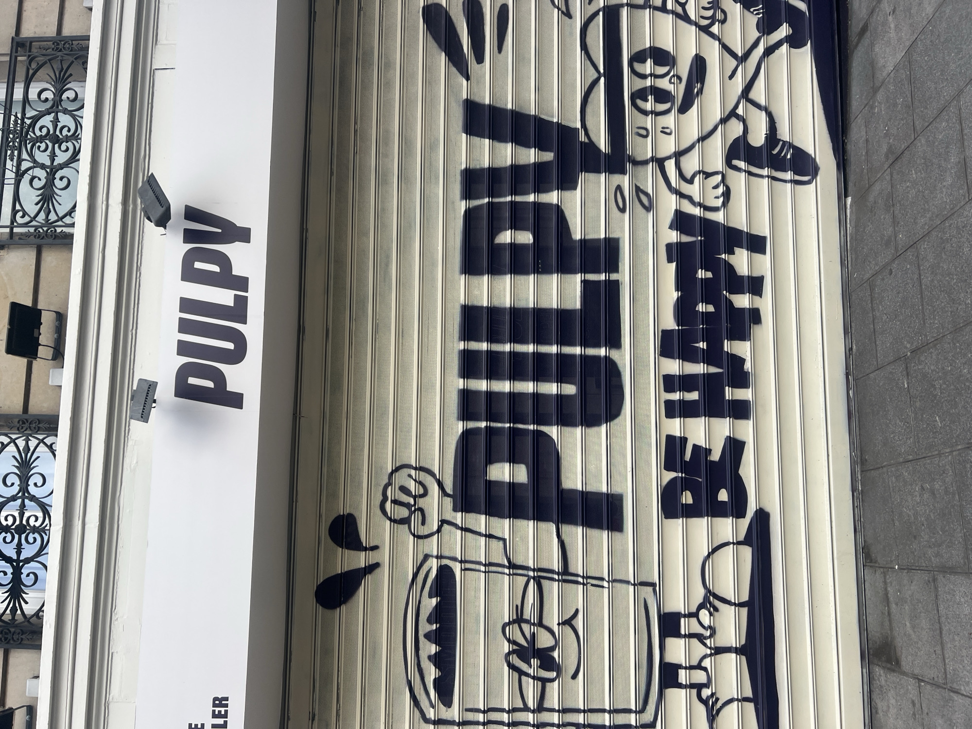 A street-level view of a closed shop front in Paris, France. The shutter is down, painted with the word "PULPY" in large, bold, dark blue letters along with cartoon-like figures and illustrations. To the left of the shutter is a vertical white sign displaying the same brand name "PULPY" in a simple, bold, dark font. A window with wrought iron detailing is visible beside the sign. The pavement is grey tiled stones.