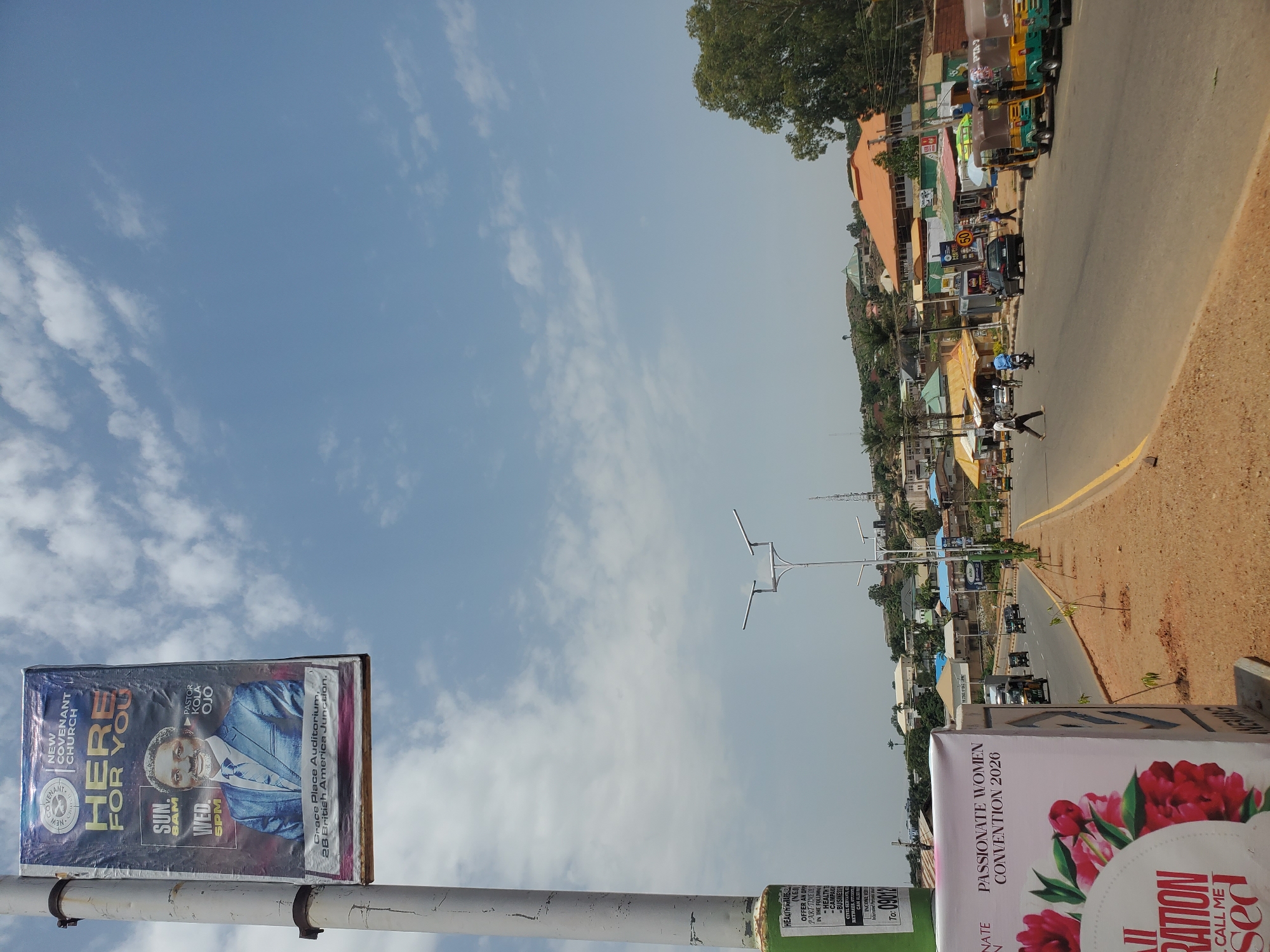 A street scene in Jos, Nigeria, is depicted under a bright, cloudy sky. A lamppost on the left displays a banner for the "New Covenant Church," featuring a portrait of Pastor Kola Ojo, service times (Sunday 8 AM, Wednesday 5 PM), and the church's location: "Grace Place Auditorium, 28 British America Junction." Below this, another banner advertises a "Passionate Women Convention 2026."
The street itself is paved, with a yellow dividing line. Several vehicles are present: a dark-colored sedan, three-wheeled auto-rickshaws (keke napep) parked on the right and in motion, and a motorcycle. Pedestrians are visible on the roadside and crossing the street. Buildings line the right side of the road, with shops and various businesses identifiable by their signage. Further in the background, hills dotted with more buildings and vegetation rise, with a prominent tower visible on one of the hilltops. The overall environment appears to be a busy urban commercial area.