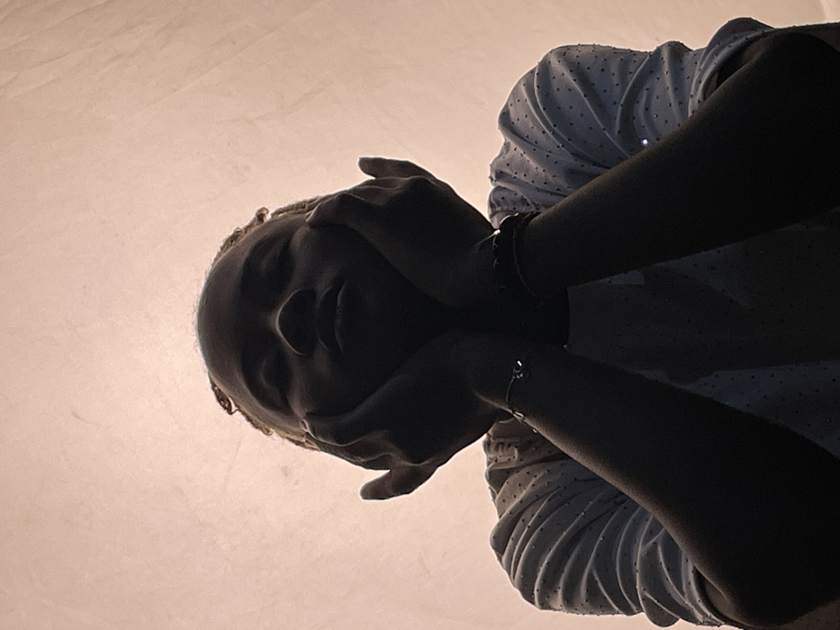 In Barakin Akawo, Nigeria, a low-angle portrait depicts an individual, likely an adult or late adolescent, from the chest up. The subject's face, hands, and forearms are largely in silhouette due to strong backlighting from a uniform, light peach-tan background. Light-colored hair, styled in small twists or braids, is visible around the upper part of the head. The subject wears a light-toned, short-sleeved garment featuring a subtle dotted texture. Both hands are raised, with palms supporting the cheeks or chin, forearms positioned vertically. On the subject's left wrist, two bracelets are present: one a slender, silver-toned chain with a small reflective charm, and the other a darker, possibly beaded or braided band. The subject's gaze is directed upwards, towards the camera. The background is a plain, subtly textured surface, indicative of an indoor or controlled setting.