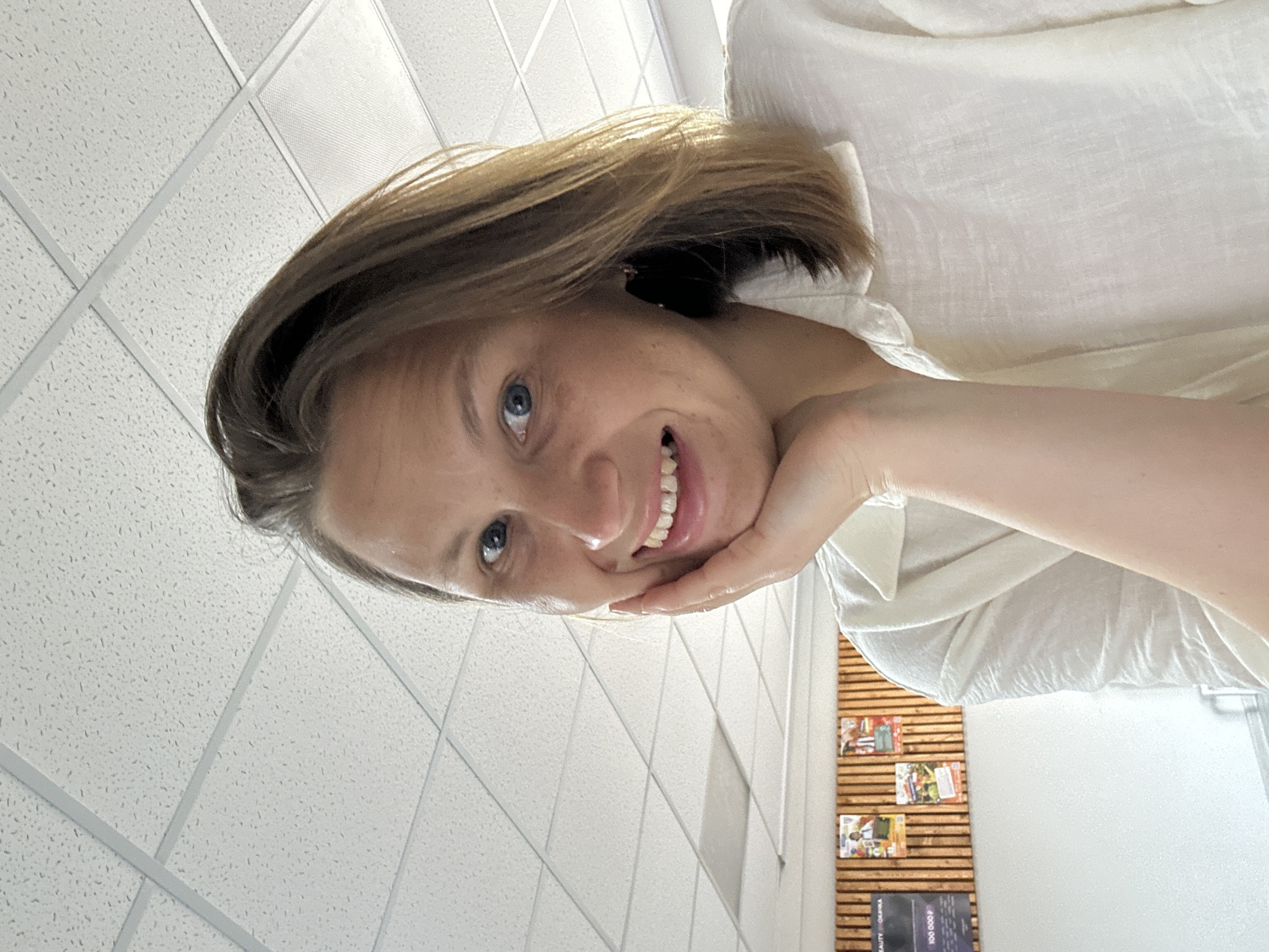 The image shows a woman with light brown hair, blue eyes, and a white shirt, resting her chin on her hand and smiling. The background appears to be an indoor space with a white tiled ceiling and a wall with wooden slats and posters or magazines. The woman is in a location that is likely in Chaikovsky, Russia.