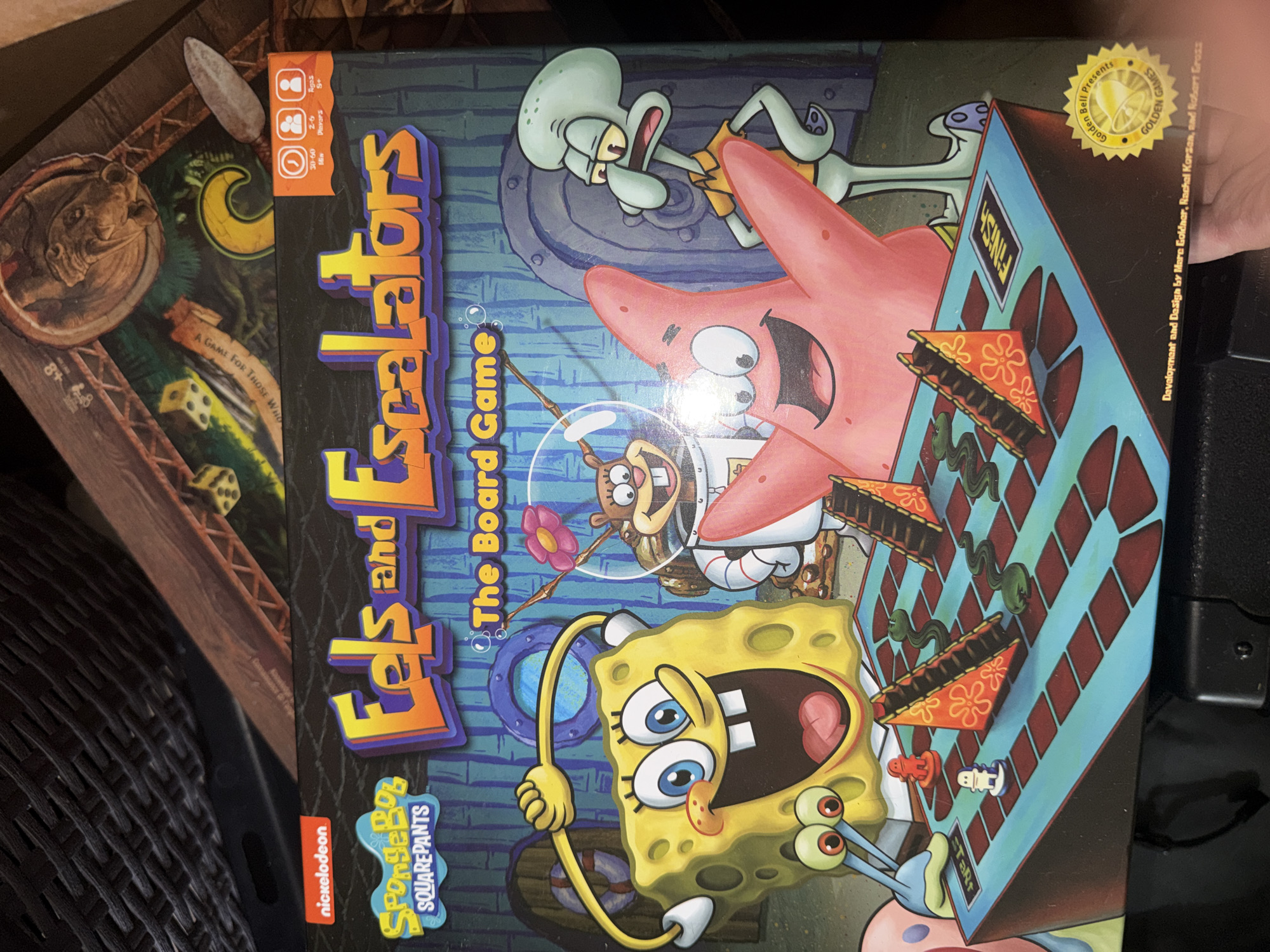 The image shows the front of a Spongebob Squarepants board game box titled "Eels and Escalators: The Board Game." The box features colorful illustrations of Spongebob, Patrick, Sandy, and Squidward. The game board itself is visible in the foreground, with Spongebob and Patrick on it, along with game pieces and dice. The box is likely displayed in a retail setting or a private collection. There is no indication of the location in Portland, United States of America.