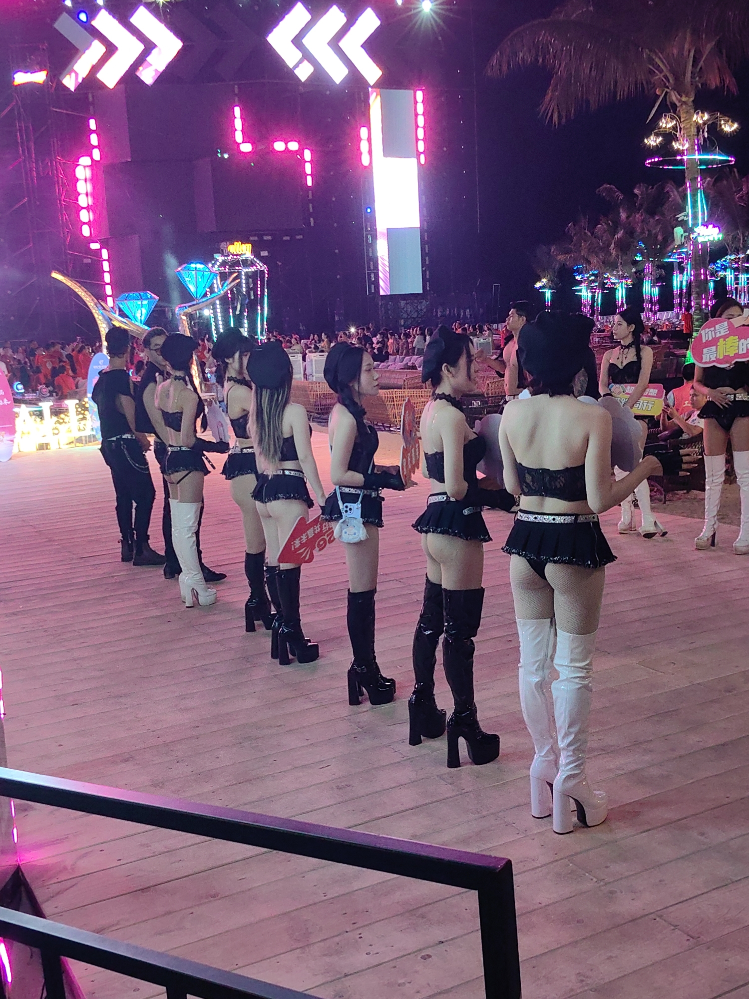 The image shows a group of women in matching outfits standing on a wooden deck in Bai Chay Ward, Vietnam. The women are wearing black tops and skirts with high white or black boots. Some are holding signs with writing in Chinese. In the background, there's a stage with bright lights and decorations, as well as trees and other structures, suggesting an outdoor event. A crowd of people can be seen in the distance. The scene is at night, and the atmosphere is festive with the illuminated stage and decorations.