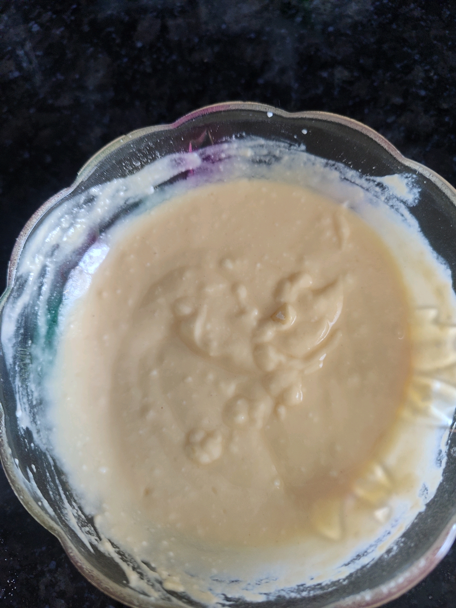 An overhead, close-up shot shows a clear glass bowl filled with a thick, pale yellow batter. The batter has a smooth, creamy consistency with a few small, lighter-colored specks scattered throughout. The bowl is placed on a dark, speckled surface, possibly a countertop. The edges of the bowl are slightly rippled. The scene is indoors, with the lighting casting subtle reflections on the batter and the bowl's surface. No people or specific environmental features of Gümüşova, Turkey are visible.