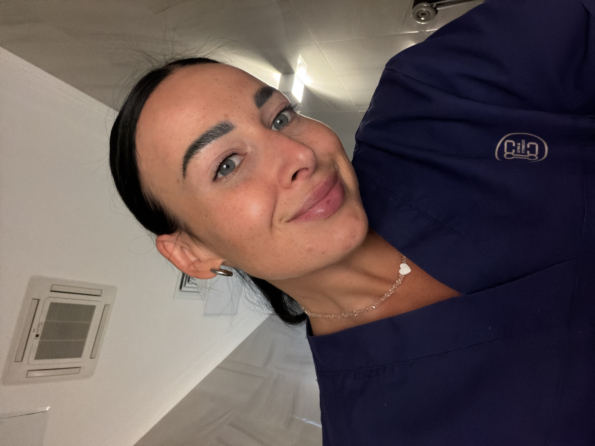 A woman is seen indoors, likely in a commercial space due to the presence of a ceiling-mounted air conditioning unit. She is wearing a dark blue shirt or uniform with a logo on the upper right, which appears to be an elliptical design containing the words "GYM & FITNESS". She also wears a delicate chain necklace with a small white heart pendant. The woman has dark hair pulled back, fair skin with freckles, and gray-blue eyes. Her expression is friendly and she appears to be smiling gently. The location, Saint Petersburg, Russia, is not discernible from the image.