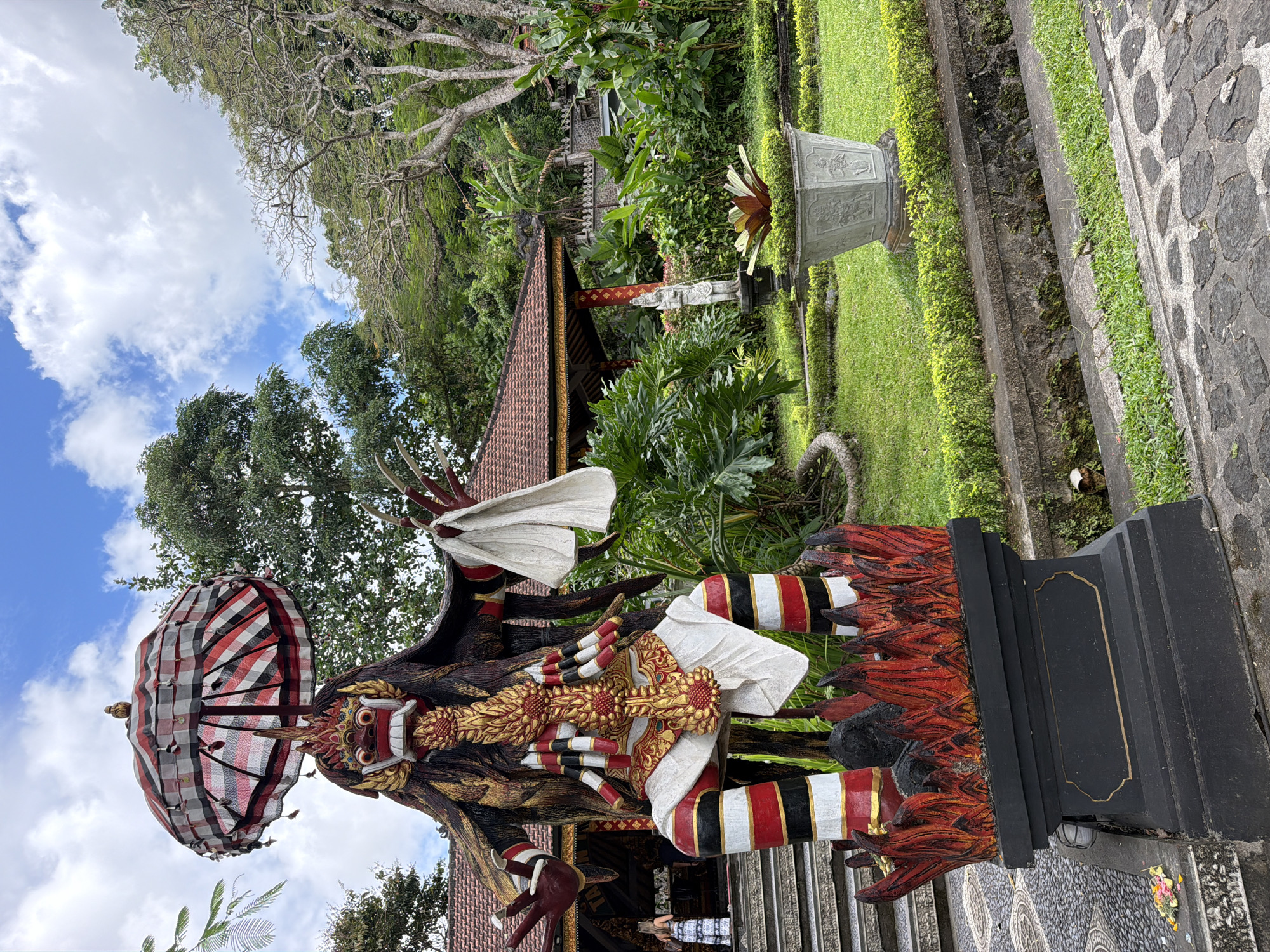 A full shot captures a large, ornate statue of a demon-like figure in the foreground, standing on a black pedestal adorned with flames. The statue is vibrantly colored with red, black, white, and gold accents, and holds a black and white checkered umbrella above its head. The figure has a fierce expression with sharp teeth and bulging eyes. Behind the statue, a traditional Balinese building with a red tile roof is visible, surrounded by lush greenery and palm trees. The scene is set in a garden with manicured lawns and stone pathways. The sky above is partly cloudy, with a mix of blue sky and white clouds. The location is the Pura Dalem Jagaraga temple in Abang, Indonesia.