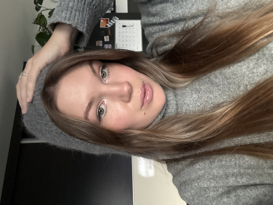 A young woman poses in a grey turtleneck sweater and fuzzy grey hat. She has long, light brown hair and light green eyes. She is facing the camera, slightly angled. Behind her is a dark monitor, a small potted plant, and a calendar. Her lips appear to be glossed. She is wearing a small ring on her left hand.