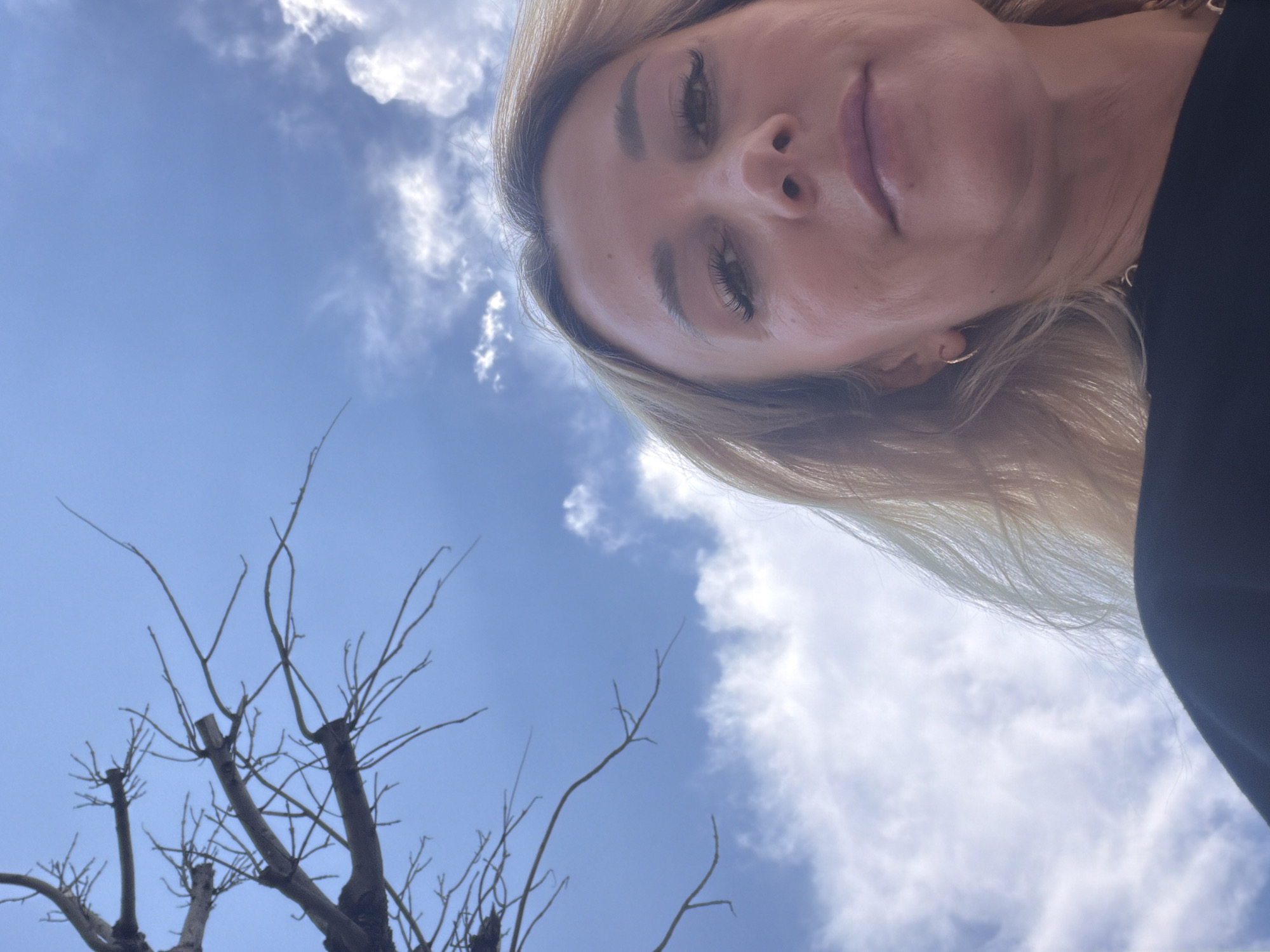 A low-angle, close-up, waist-up shot captures a woman with blonde hair, light skin, and makeup. She is wearing a black top. Her face is turned towards the camera, with her eyes looking forward. A bare tree branch extends from the upper left corner of the frame, against a bright blue sky with scattered white clouds. The scene appears to be outdoors in Kemer, Turkey, based on the hint provided.
