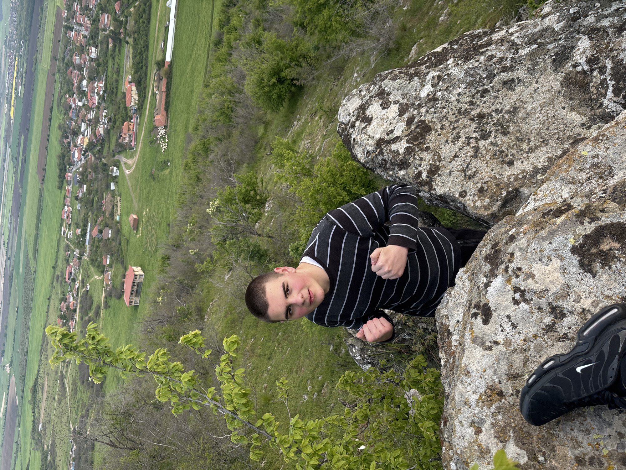 A young person stands on a mossy rocky ledge in Goritsa, Bulgaria, looking down at the town below. The town is located in a valley with green fields and scattered buildings. The person is wearing a black and white striped shirt and black shoes.