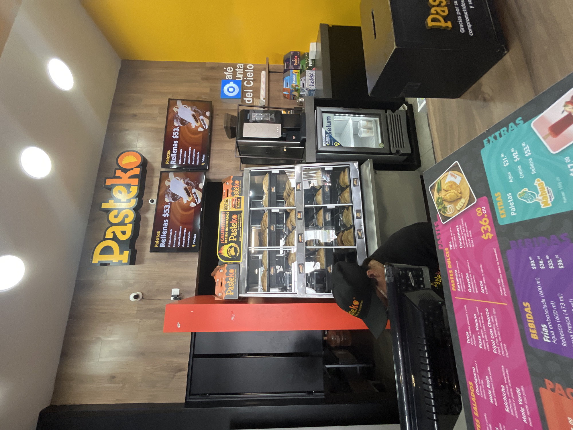 The image depicts the interior of a "Pasteko" establishment, likely in Puebla, Mexico, a local franchise specializing in savory and sweet pastries ("pastes"). The scene includes:
**Environment:** The location features wood-paneled walls with yellow accent paint near the ceiling. There's a display case showcasing the "pastes", a beverage cooler, and menus posted on the wall and at the counter.
**Objects:** Display cases filled with pastries, a beverage cooler, printed menus advertising different "paste" varieties and their prices, "Pasteko" signage, a coffee machine, and a donation box.
**People:** One person with a "Pasteko"-branded hat. They appear to be an employee working behind the service counter, attending to the pastry display or possibly serving customers.