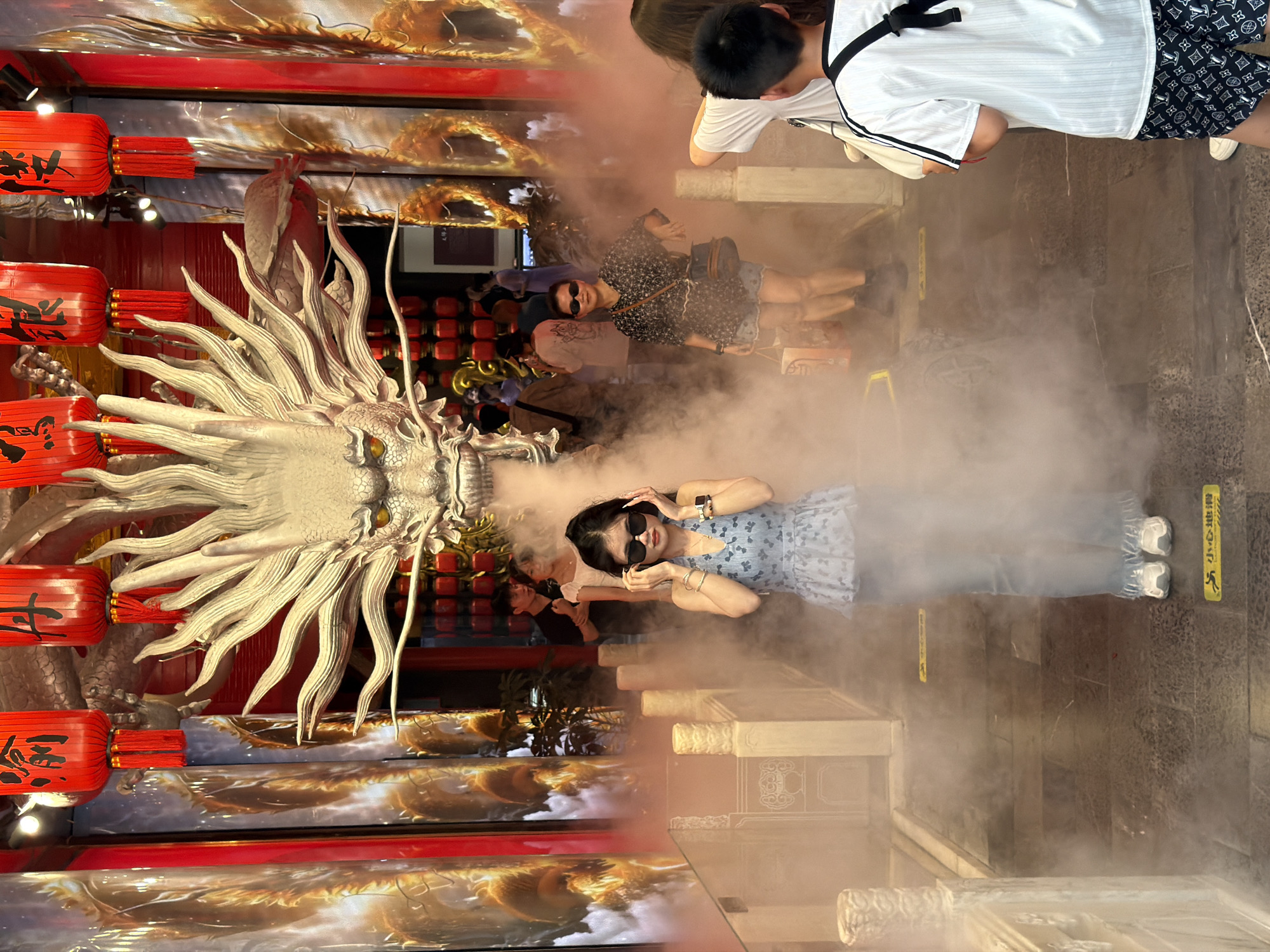 The image captures an outdoor scene at what appears to be a cultural attraction, possibly in Yuzhong District, China, given the prominent dragon motif and traditional red lanterns. In the foreground, a young woman stands amidst a haze of what seems to be dry ice or smoke, adjusting her sunglasses. She is wearing a light blue patterned top and light-colored pants. Behind her, a large, ornate dragon head sculpture dominates the background, with red lanterns hanging above. Several other individuals are visible in the mid-ground and background, some appearing to be tourists or visitors. The environment suggests an urban setting with architectural elements that hint at traditional Chinese design, including what looks like balustrades and decorative pillars. The lighting is moderate, with the red lanterns providing a splash of color.