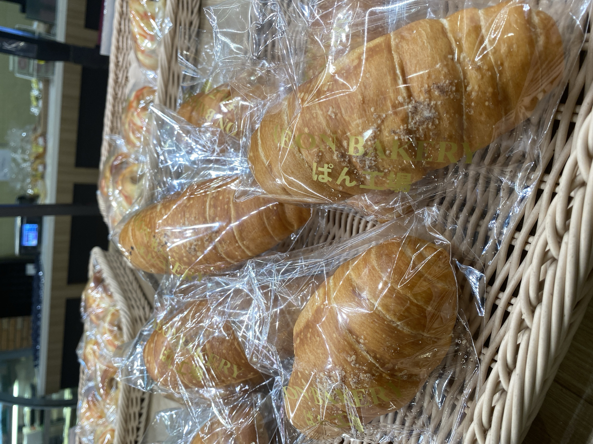 The image shows a close-up view of freshly baked bread products displayed in woven baskets at a bakery. The bread items, predominantly long rolls and croissants, are individually wrapped in clear plastic packaging. Some of the rolls have a golden-brown crust and are sprinkled with sugar or other toppings. The background suggests an indoor setting, possibly a retail environment with shelves and a counter visible. The lighting is bright, highlighting the texture and color of the baked goods. The overall scene conveys a sense of a well-stocked bakery offering a variety of pastries. The specific location in East Jakarta, Indonesia is not discernible from the visual information provided in the image.