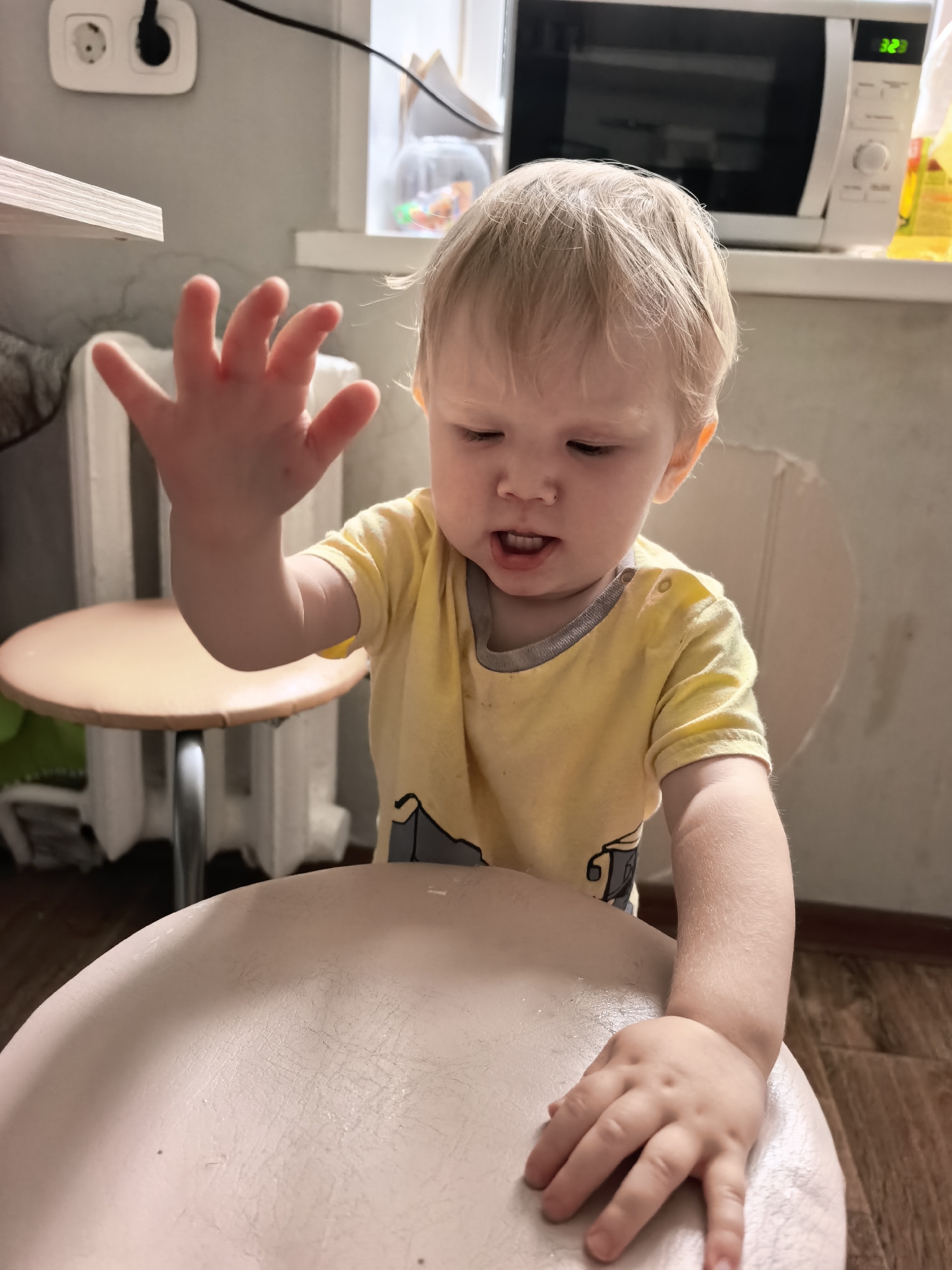 A toddler with blonde hair and blue eyes, wearing a yellow t-shirt, is captured in a close-up indoor shot. The toddler's right hand is raised, fingers spread, while their left hand rests on a light-colored surface, likely a chair or table. The background reveals a microwave, a radiator, and what appears to be a window with some items inside. The lighting suggests an interior setting, possibly a kitchen or dining area. The scene is devoid of any specific identifying markers that would pinpoint its location within Sterlitamak, Russia.