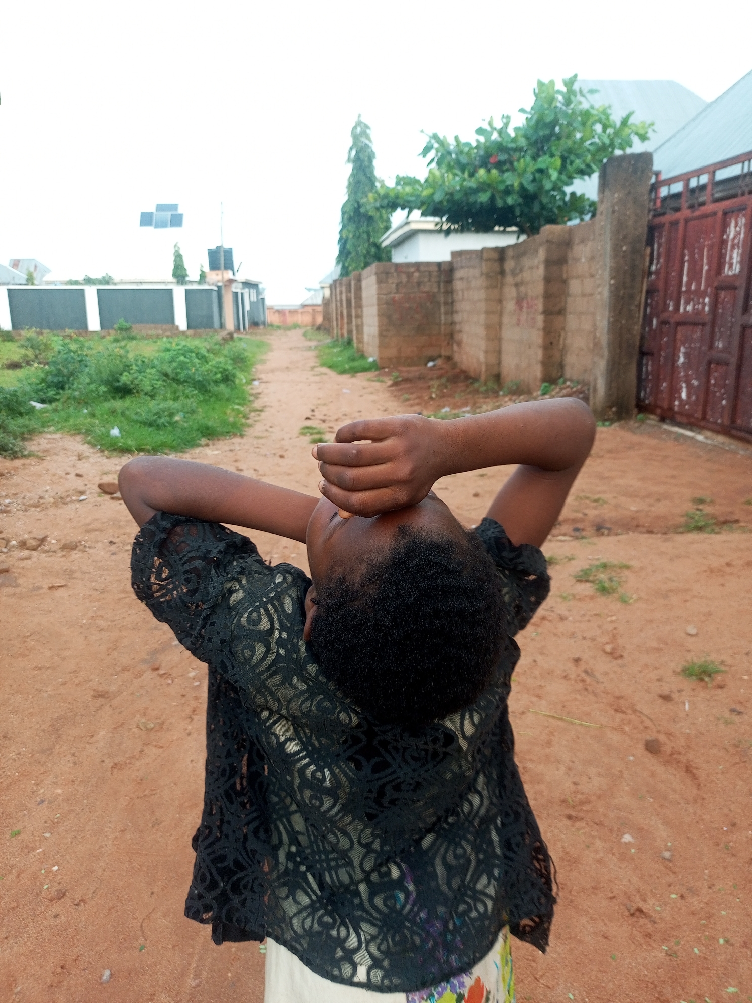 A young girl stands in a sandy, unpaved alleyway in Tempure, Nigeria. She is wearing a black lace top over a colorful patterned skirt. Her arms are raised, with her hands covering her face, as if shielding her eyes or expressing distress. The alleyway is lined with sparse vegetation and brick walls, with buildings and structures visible in the background. A large, red metal gate is prominent on the right side of the image. The sky is overcast and bright.