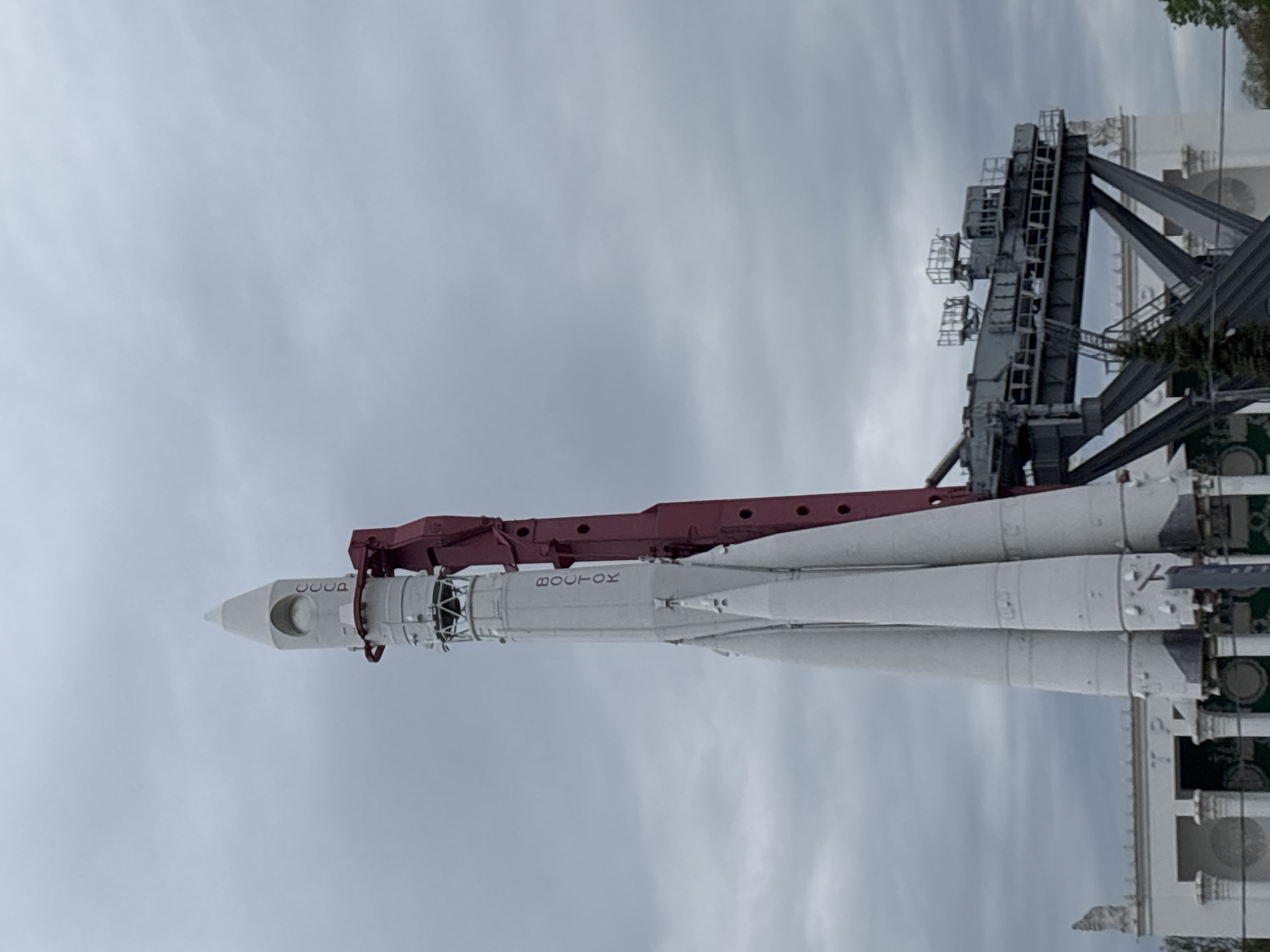 The image shows a Vostok rocket on display at the Exhibition of Achievements of National Economy (VDNKh) in Moscow, Russia. The rocket is white with red markings and is positioned on a launch platform against a cloudy sky. The rocket is oriented vertically, with its nose cone pointing upwards. The inscription "CCCP" (USSR) and "VOSTOK" are visible on the side of the rocket. A gray metal launch support structure with several levels is next to the rocket. The base of a white building with ornate details is partially visible behind the launch structure.