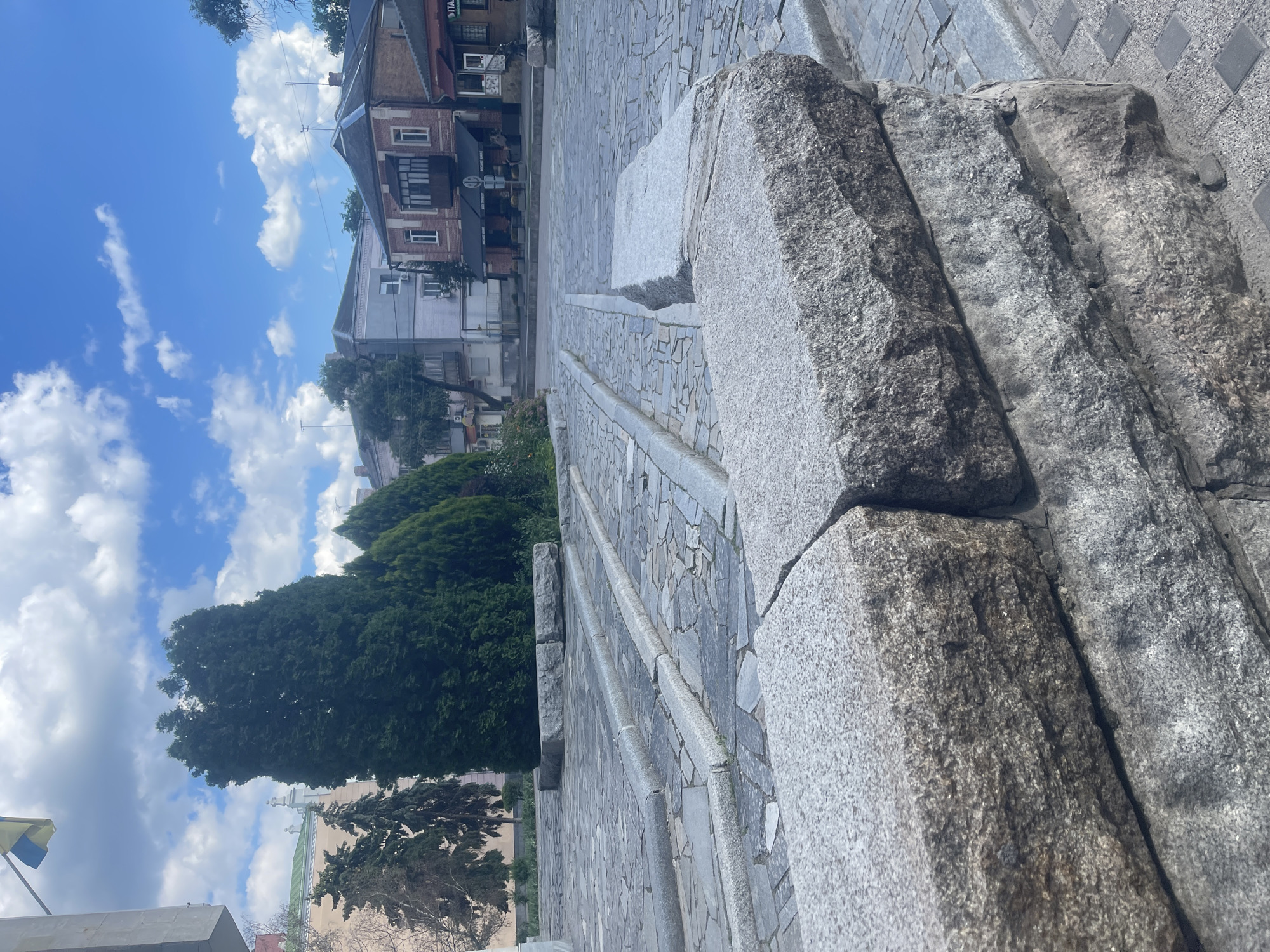 The image shows a paved public space with stone elements in Poltava, Ukraine. In the foreground, large, rough-hewn granite blocks form a low wall or platform. The paving stones are a mosaic of grey, irregularly shaped pieces, with some areas featuring raised linear stone borders. In the midground, a row of tall, dark green coniferous trees stands behind the stone features, suggesting a park or garden area. Further back, a street scene unfolds with buildings of various architectural styles, including brick and stucco structures, some with balconies and awnings. A Ukrainian flag is visible flying on a pole in the upper left corner of the image. The sky is blue with scattered white clouds. The scene appears to be a public square or walkway, likely in an urban setting.