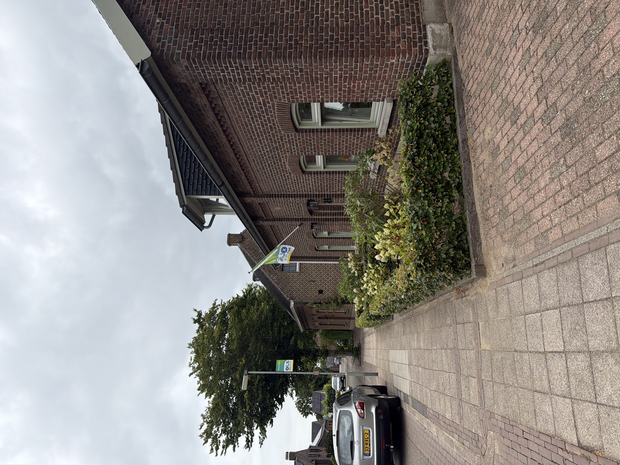 The image depicts a street scene in Melick, Netherlands, on an overcast day. In the foreground, a silver Nissan Qashqai with license plate 33-RXL-2 is parked on the left side of the street. To the right of the car, a paved sidewalk runs along a brick building. A street lamp with a sign that reads "OLS MELICK" is positioned next to the sidewalk.
The building on the right is constructed of red brick and features multiple windows with light green frames. A green and white flag is flying from a flagpole attached to the building. In front of the building, a well-maintained hedge with flowering plants forms a border along the sidewalk.
The street is paved with reddish-brown bricks, and the sidewalk is composed of grey concrete pavers with some brick detailing. In the background, more houses and trees can be seen, and the sky is heavily clouded. The scene appears to be a quiet residential street.