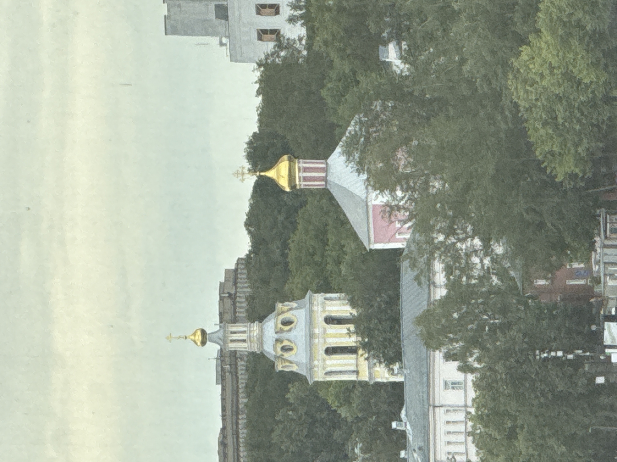 This is a photograph of the city of Moscow, Russia. The image depicts a cityscape with several buildings and trees. In the foreground, a light-colored building with multiple windows is visible amongst lush green trees. To the left, a tall, slender yellow tower with a grey roof and a golden dome stands out against the sky. Further to the right, another building with a pink facade and a grey roof topped with a golden dome and cross is partially obscured by trees. In the background, a large, multi-story grey building with visible windows can be seen. The sky is overcast with a pale, hazy appearance.