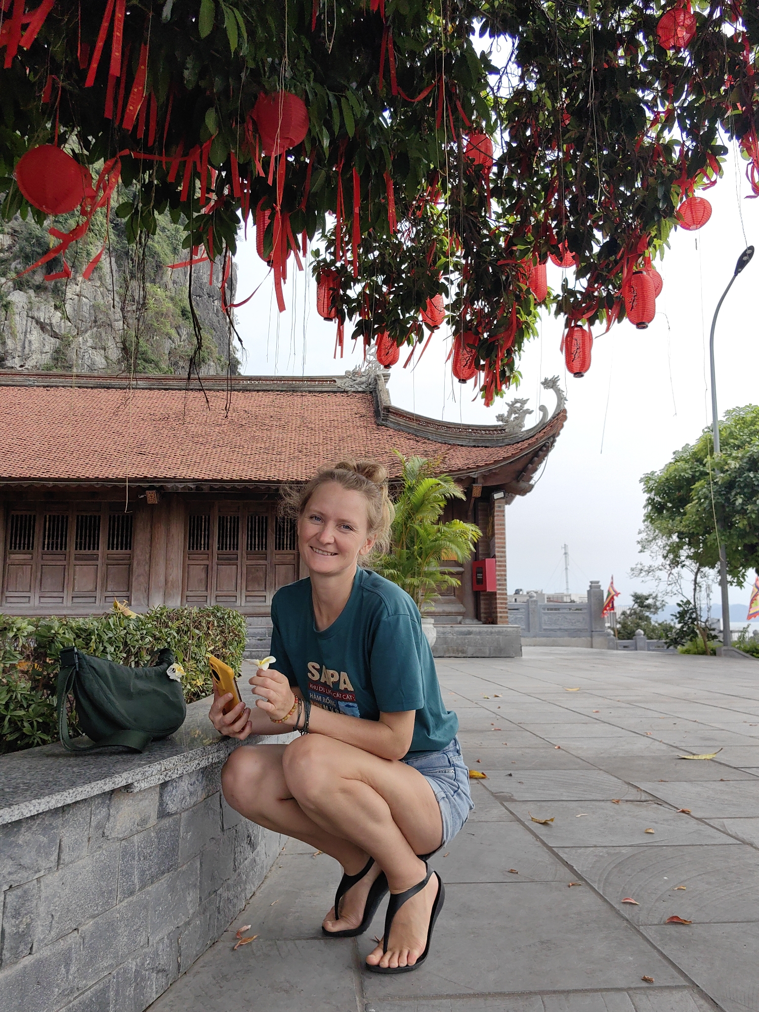 A smiling woman with blondish hair tied in a bun is captured crouching outdoors in what appears to be a temple or pagoda complex in Hong Gai Ward, Vietnam. She is wearing a dark teal t-shirt, denim shorts, and black flip-flops. In her left hand, she holds a yellow smartphone, and in her right, a small white flower, likely a frangipani. A green shoulder bag rests on a low stone wall beside her.
Above her and extending across the upper portion of the frame is a large tree with dense green foliage, heavily decorated with numerous red paper lanterns and long red ribbons, creating a vibrant canopy. In the background, a traditional Vietnamese building with an ornate reddish-brown tiled roof and dark wooden doors is visible. Behind the building, rugged limestone karsts characteristic of the region rise under a bright, overcast sky. The ground is paved with large grey tiles, scattered with a few fallen leaves.
On the woman's t-shirt, the word "SAPA" is prominently displayed, along with smaller Vietnamese text below it: "KHU DU LỊCH CÁT CÁT" (Cat Cat Tourist Area) and "HÀM RỒNG" (Ham Rong – Dragon Jaw Mountain), referring to well-known locations in Sapa, Vietnam. Her joyful expression suggests she is having a pleasant time. The scene is illuminated by diffuse daylight, indicative of an overcast day.