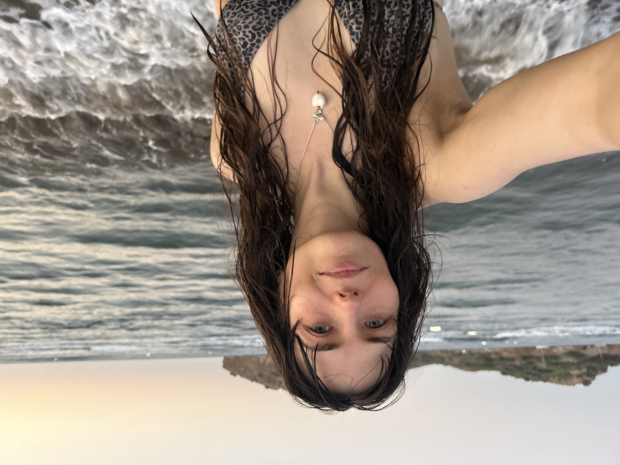 A young woman with dark, wet hair stands on a beach in Ortaca, Turkey, at dusk. She is wearing a leopard-print bikini and a silver necklace with a heart-shaped pendant. The ocean is behind her, with waves crashing on the shore. In the distance, a small island with vegetation is visible. The sky is a soft gradient of orange and gray.