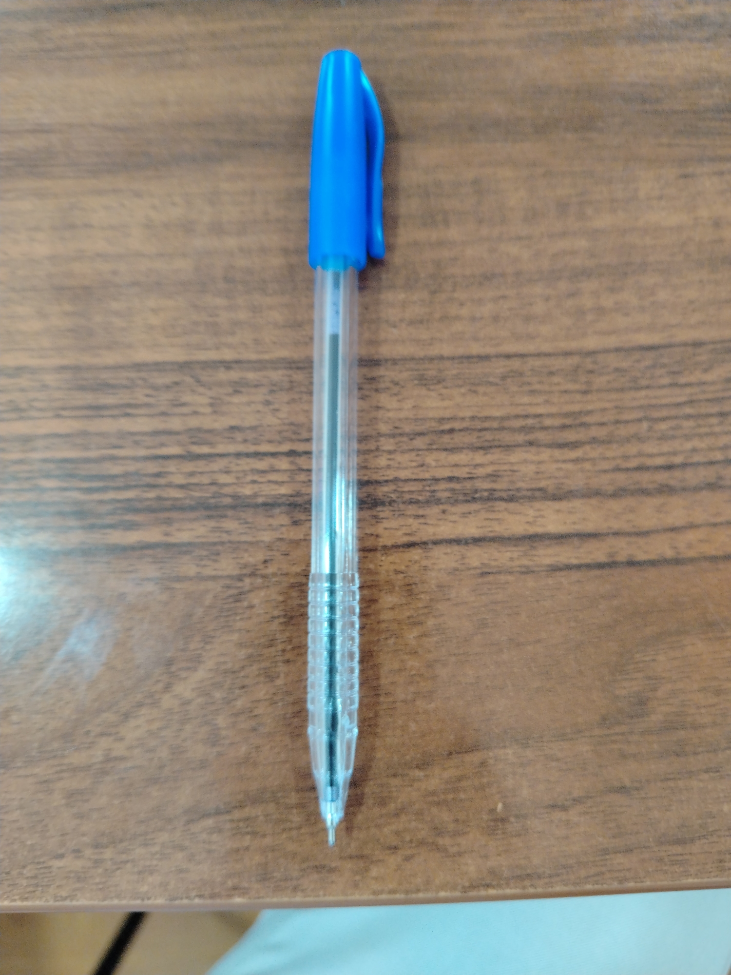 A blue pen with a clear plastic barrel is lying on a brown wooden surface. The pen is positioned vertically in the center of the image, with its cap at the top and the tip pointing downwards. The wooden surface has a visible grain pattern. This image does not contain information about the city of Nurata, Uzbekistan.