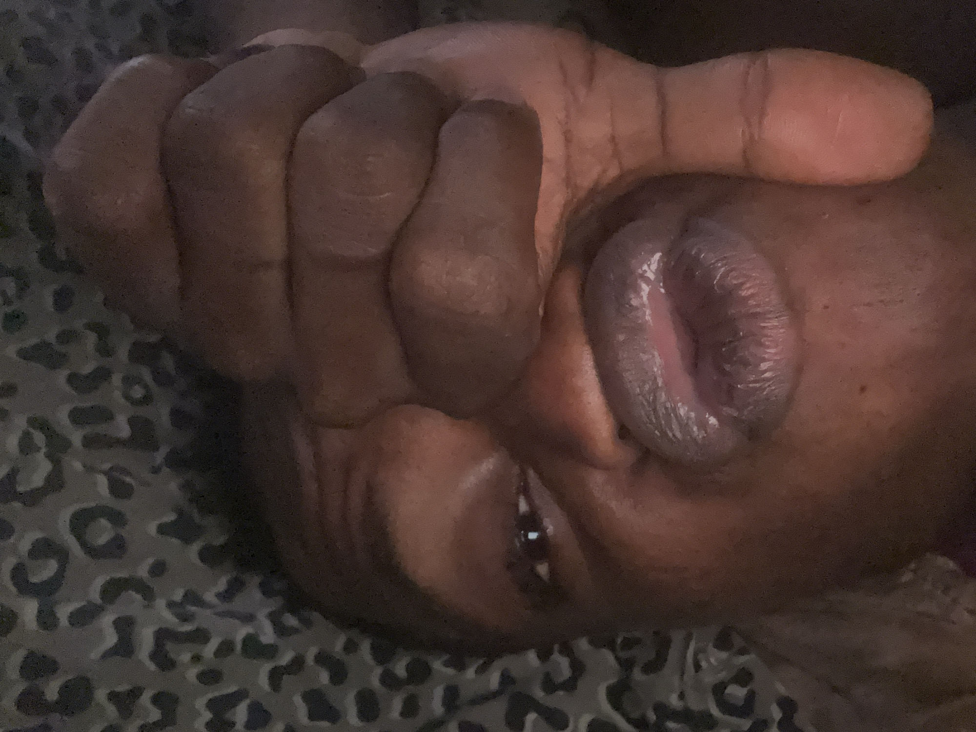 The image is a close-up, low-angle, indoor shot of a person's face, obscured by their hand. The person has dark skin and is wearing lip gloss. Their hand is positioned in front of their face, obscuring most of their features. The background is a patterned fabric, possibly a blanket or upholstery, with a leopard-like print in shades of grey and black. The lighting is dim, creating shadows and highlighting the texture of the skin and lips. There are no visible elements that would suggest a specific location within Ikorodu, Nigeria.