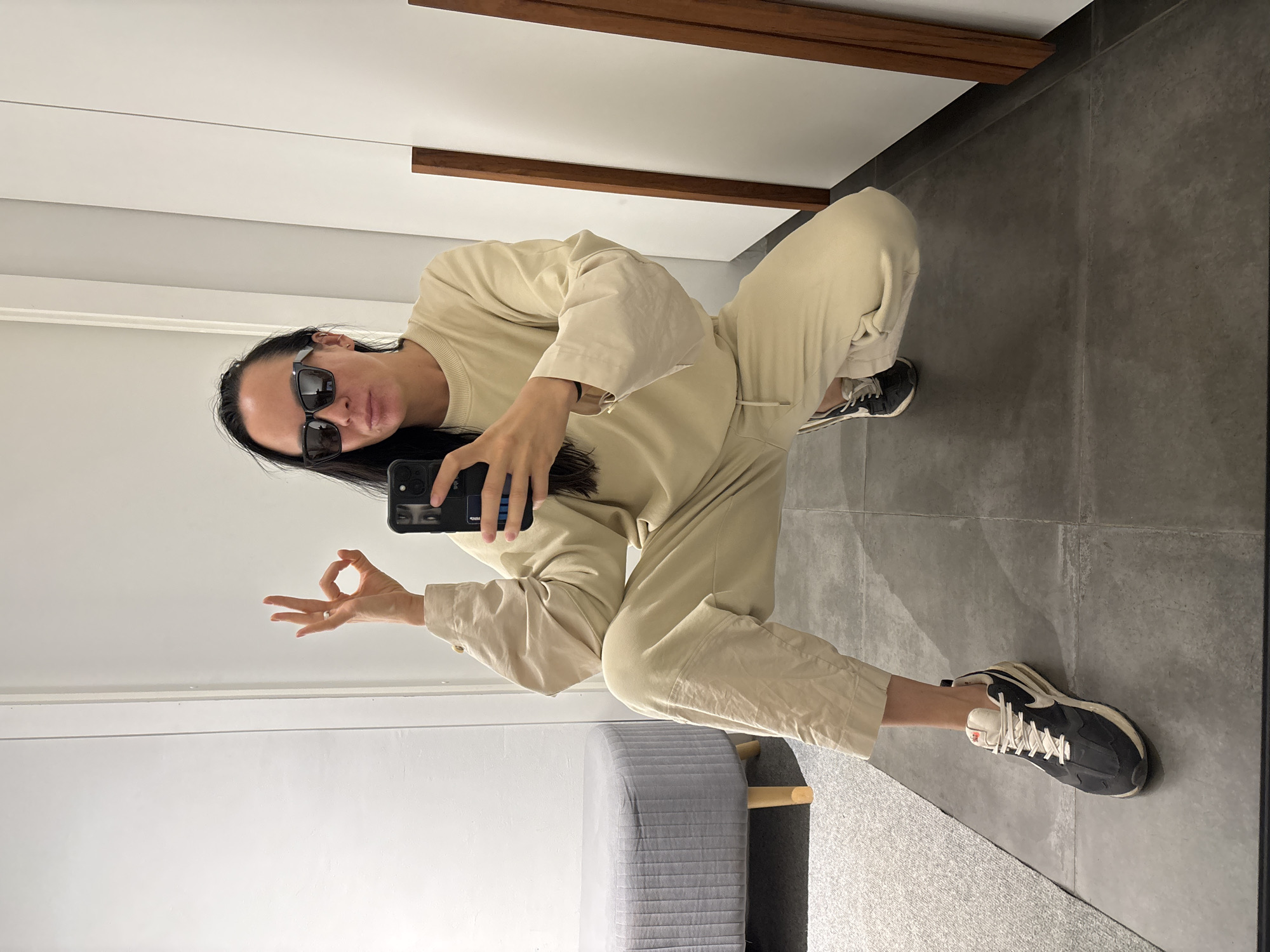 A person is crouching in a brightly lit room, taking a selfie in a mirror. The person is wearing sunglasses and a matching beige sweatsuit and is holding a phone in their left hand while making an "OK" gesture with their right hand. The phone has a black case with a design featuring eyes. The person has long dark hair. The floor is gray tile. In the background are white walls with wood trim, a gray upholstered stool, and a glimpse of a doorway. The city of Chaikovsky, Russia is not identifiable from this image.