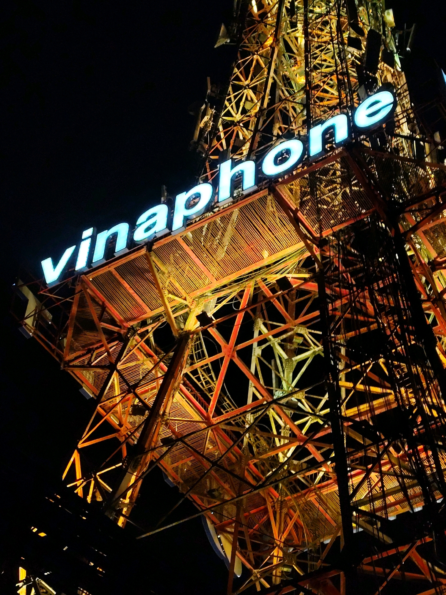 A nighttime, low-angle shot captures the top section of the Vinaphone telecommunications tower in Da Lat, Vietnam. The tower, made of a crisscrossing metal structure, is illuminated with a warm orange glow from internal lights, creating a striking contrast against the dark sky. A prominent, illuminated sign spelling out "vinaphone" in bright blue letters is affixed to the tower's upper section. The sign is sharp and clear, with each letter glowing with an ethereal light. The metallic structure of the tower is intricate and geometric, with many beams and supports visible. The scene is devoid of people or any discernible activity, focusing solely on the architectural and signage elements of the tower.