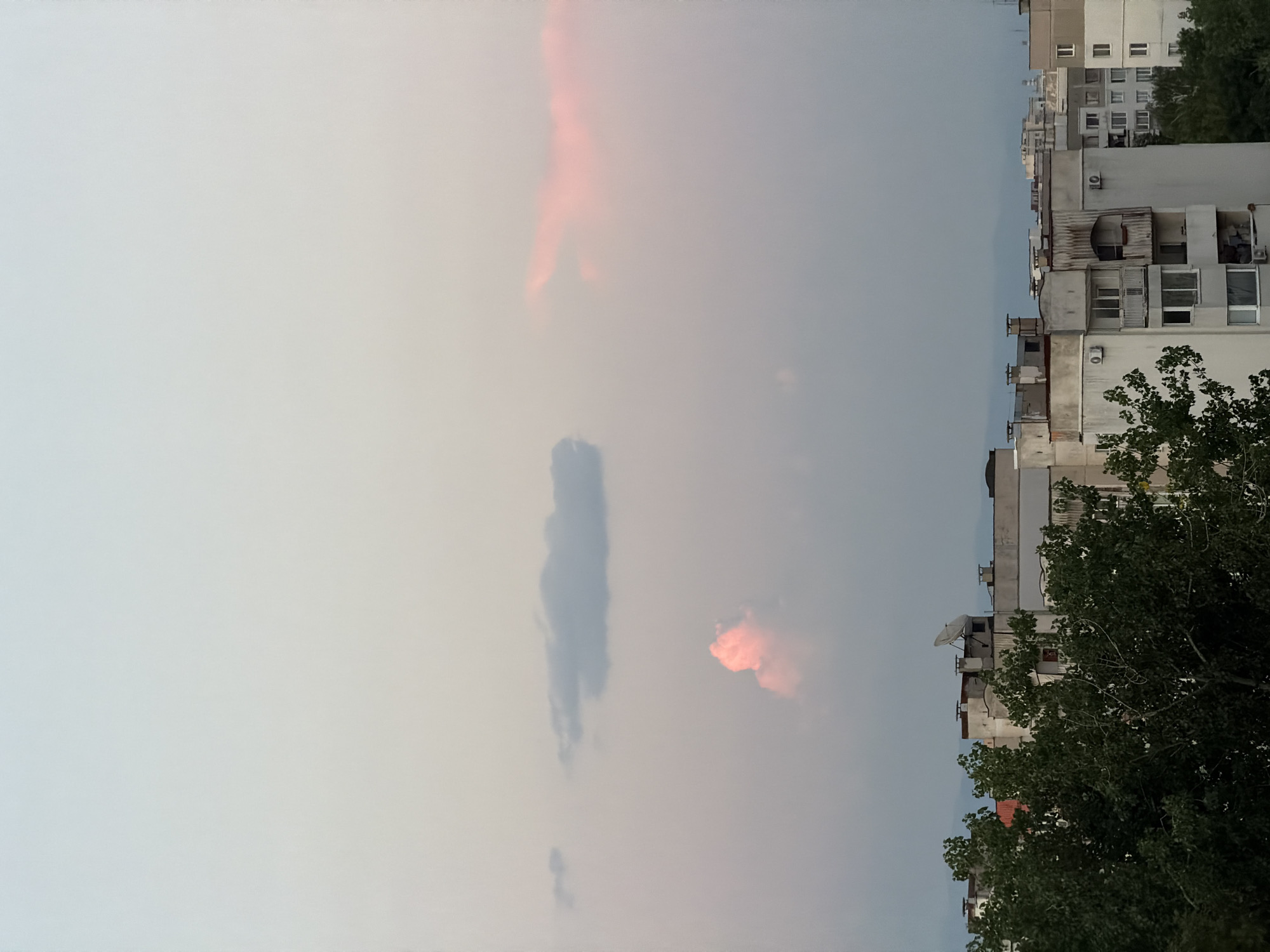 This is a vertical, eye-level, outdoor, daytime photograph taken in the city of Sofia, Bulgaria, likely during twilight. The scene is dominated by a pale sky with subtle hues of blue, gray, and a hint of pink from the setting sun. Two distinct cloud formations are visible: a long, thin gray cloud stretches across the upper middle portion of the frame, and a smaller, puffy cloud with a bright pink glow is positioned below and to the left of it. In the foreground, the tops of several apartment buildings are visible, characterized by their concrete facades and numerous windows. Trees with dark green foliage partially obscure the lower sections of these buildings. The architectural style suggests a Soviet-era residential block. There are no people visible in the frame. The overall atmosphere is calm and serene, capturing the transition from day to night over an urban landscape.