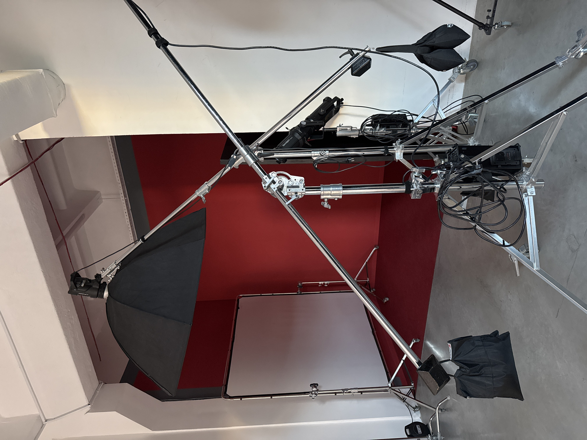 The image depicts a photography studio setup indoors. The walls are painted white and a portion is covered in red. The floor is concrete. Lighting equipment includes a large softbox on a stand and what appears to be a boom arm supporting lights or modifiers above. There is also a backdrop frame holding a white scrim. Cables and wires are visibly connected to the equipment. It is not possible to determine the location in Minsk, Belarus from the image alone.