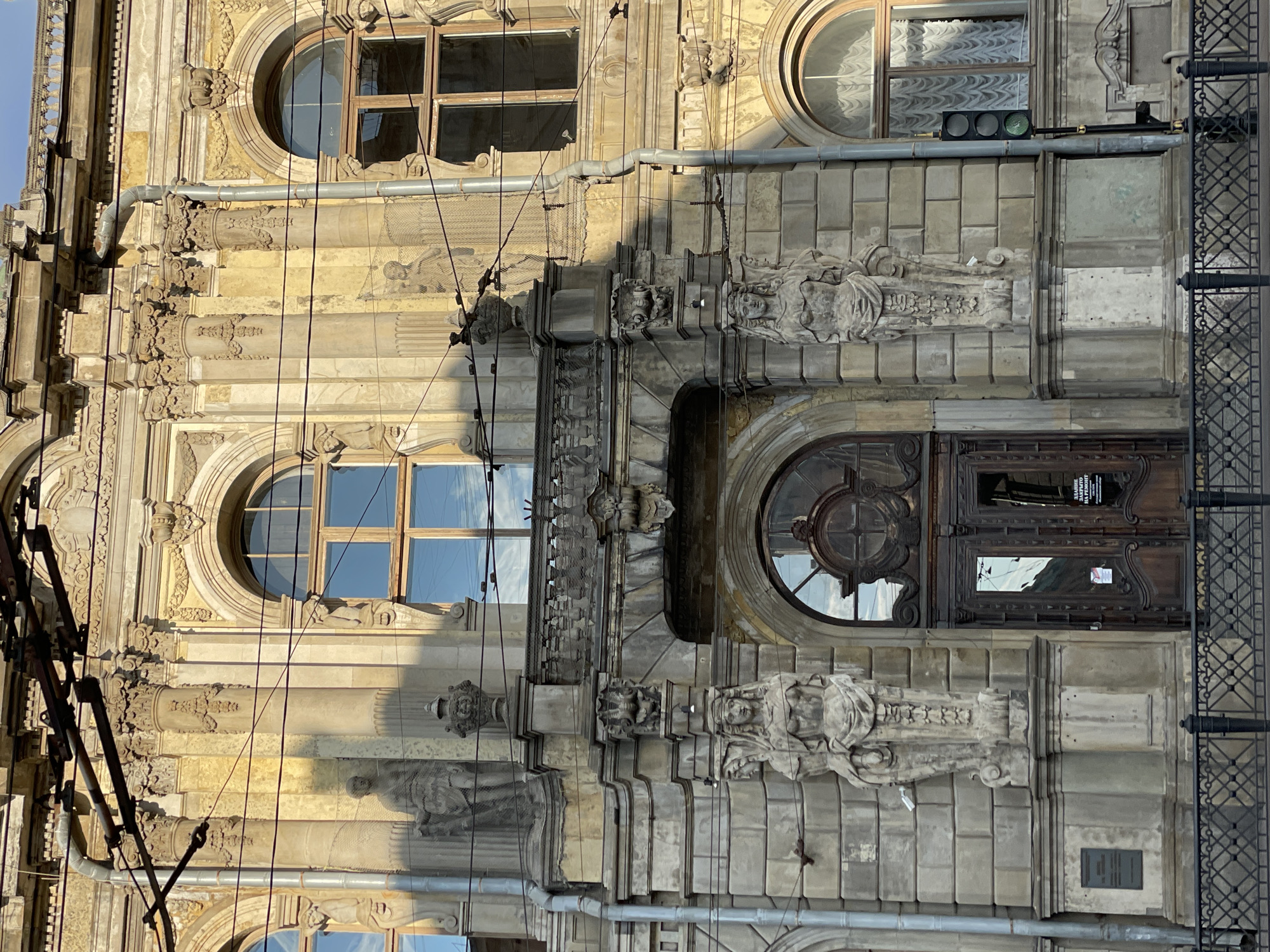 The image presents a portion of an ornate facade in Saint Petersburg, Russia, showcasing architectural elements indicative of a classical or baroque revival style. The building features a large arched doorway with carved wooden doors. Flanking the entrance are sculpted figures, possibly caryatids or allegorical representations, integrated into the stone facade. Above the entrance are arched windows. A network of utility lines runs across the front of the building, juxtaposing modern infrastructure with the historical architecture. The building appears to be constructed from stone or concrete, with a weathered texture. A metal railing is seen along the right side of the image. The overall impression is one of aging grandeur in an urban setting.