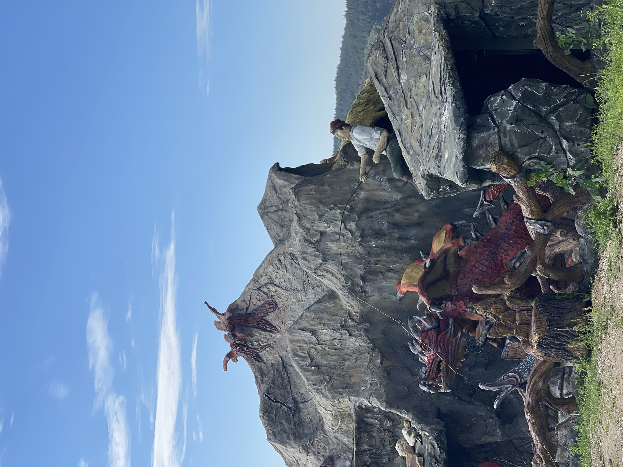 This image captures an outdoor, mythological-themed art installation or attraction situated in Koprinka, Bulgaria, under a bright blue sky with scattered white cirrocumulus clouds. The central feature is a large, multi-tiered artificial rock formation resembling a mountain or cliff face.
On the upper right side of the formation, a life-sized statue of a man is depicted sitting on a rocky ledge. The man wears a light-colored long-sleeved shirt, dark pants, and a reddish-brown cap, and is actively holding a fishing rod, with the line extending downwards towards the base of the structure. His posture suggests he is straining or pulling on the line.
At the base of the main rock formation, partially obscured by the foreground and artificial rockwork, is a large, detailed statue of a red dragon with prominent yellow and orange wings. The dragon's mouth is open, exposing teeth, and it appears to be caught or interacting with the fishing line cast by the man above. Adjacent to the dragon on the left, another creature with an open, elongated mouth emerges from a darker recess, and various sculpted wooden or root-like elements are positioned around the dragon. On the highest peak of the artificial mountain, a dark reddish-brown, gnarled structure resembling roots or tentacles protrudes upwards.
In the far background on the right, a line of dark green forested hills extends horizontally beneath the clear sky. The ground in the immediate foreground consists of patches of green grass interspersed with exposed dirt or gravel.