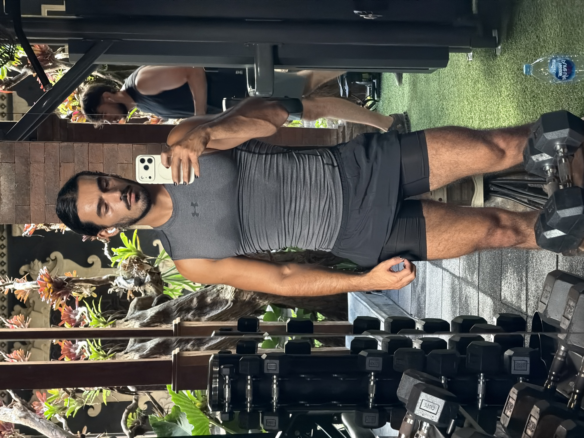In the city of North Kuta, Indonesia, a man of Middle Eastern descent is taking a mirror selfie in a gym. He is wearing a gray Under Armour sleeveless shirt and black athletic shorts. The man has dark hair tied back in a bun and a beard. He is holding a white iPhone in his right hand. In the background, other gym equipment, including dumbbells, are visible. There is also another man in the background wearing a black sleeveless shirt. The gym appears to be well-lit and decorated with plants and wooden elements.
