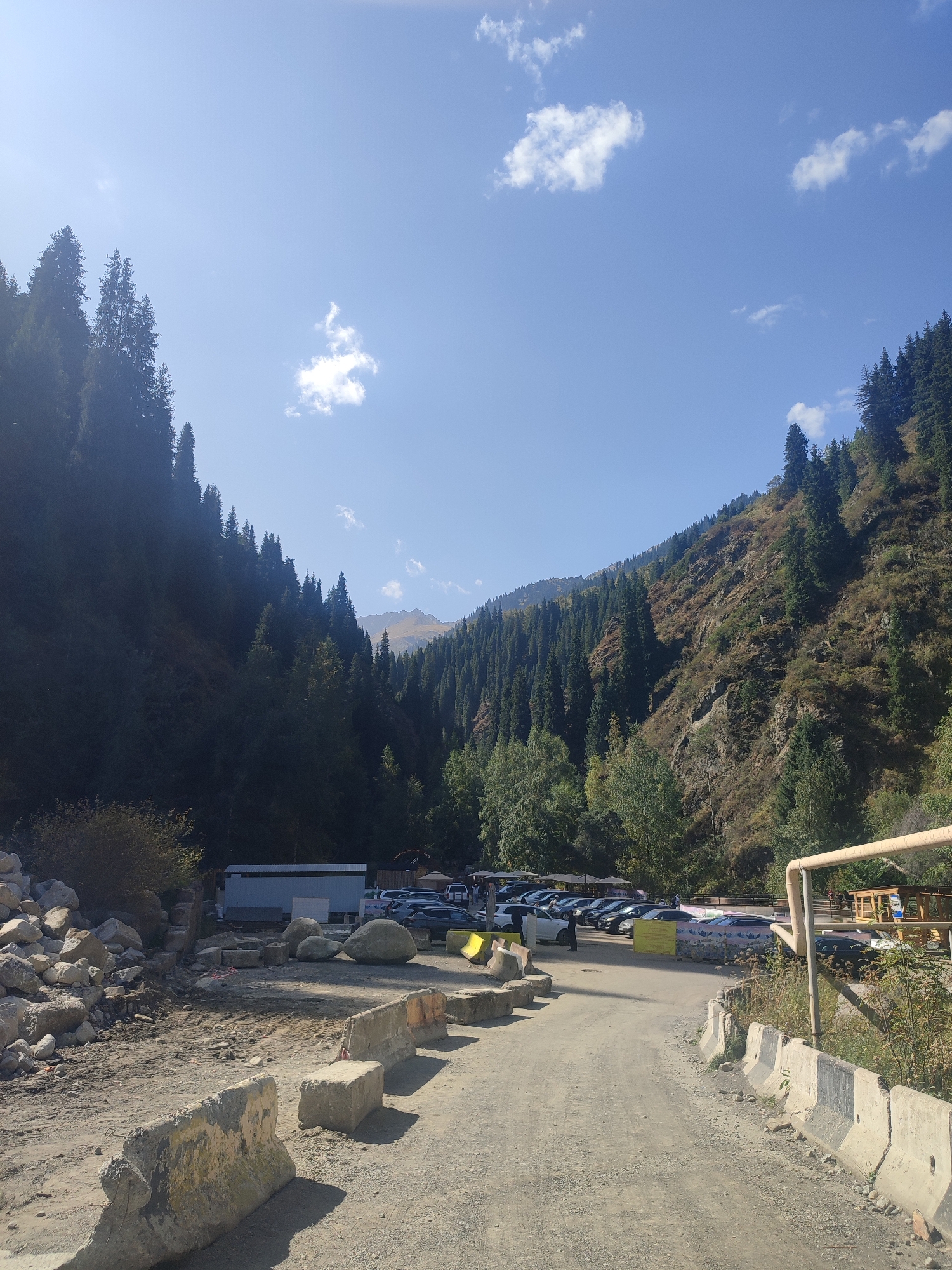 The image shows a mountainous landscape with a road leading into a valley. The valley is densely forested with evergreen trees on both sides, with steeper, rocky slopes on the right side of the frame. The sky is clear blue with a few scattered white clouds. In the foreground, there is a construction or road maintenance area with large rocks, concrete barriers, and a dirt road. A small building or temporary structure is visible on the left side of the road, behind the rocks. Further down the road, a parking lot with several cars is visible, along with some wooden structures and people milling about. The scene appears to be a natural area on the outskirts of Almaty, Kazakhstan, likely a popular tourist or recreational destination given the presence of a parking lot and amenities. The overall impression is one of natural beauty with some ongoing infrastructure work.