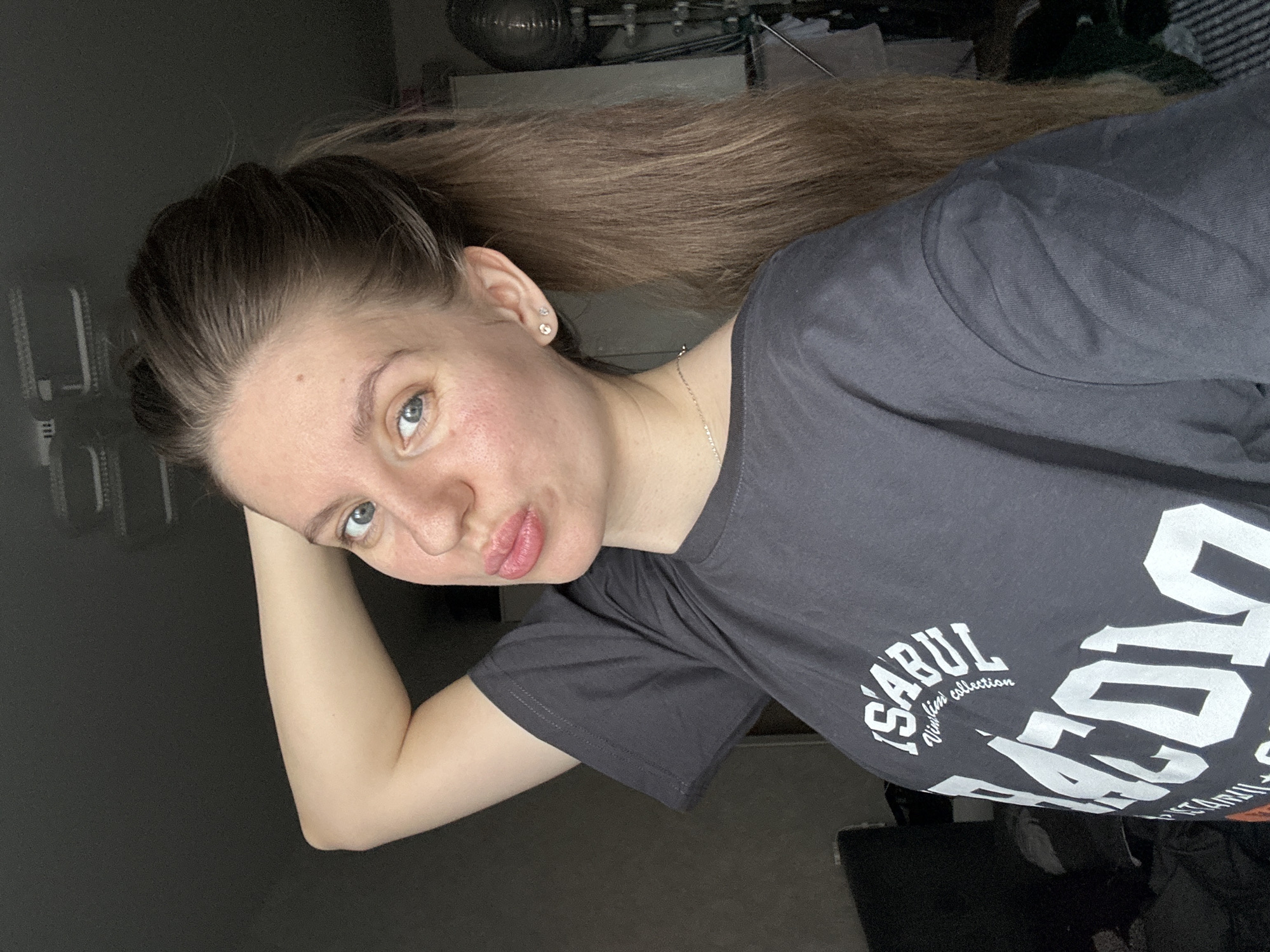 A young woman with blonde hair in a high ponytail is posing for a selfie. She is wearing a gray t-shirt with white text that reads "İstanbul new lim collection." She has fair skin and blue eyes. The background is blurred, but it appears to be an indoor setting with some objects visible. The location is not identifiable as the city of Klin, Russia, from the provided image.