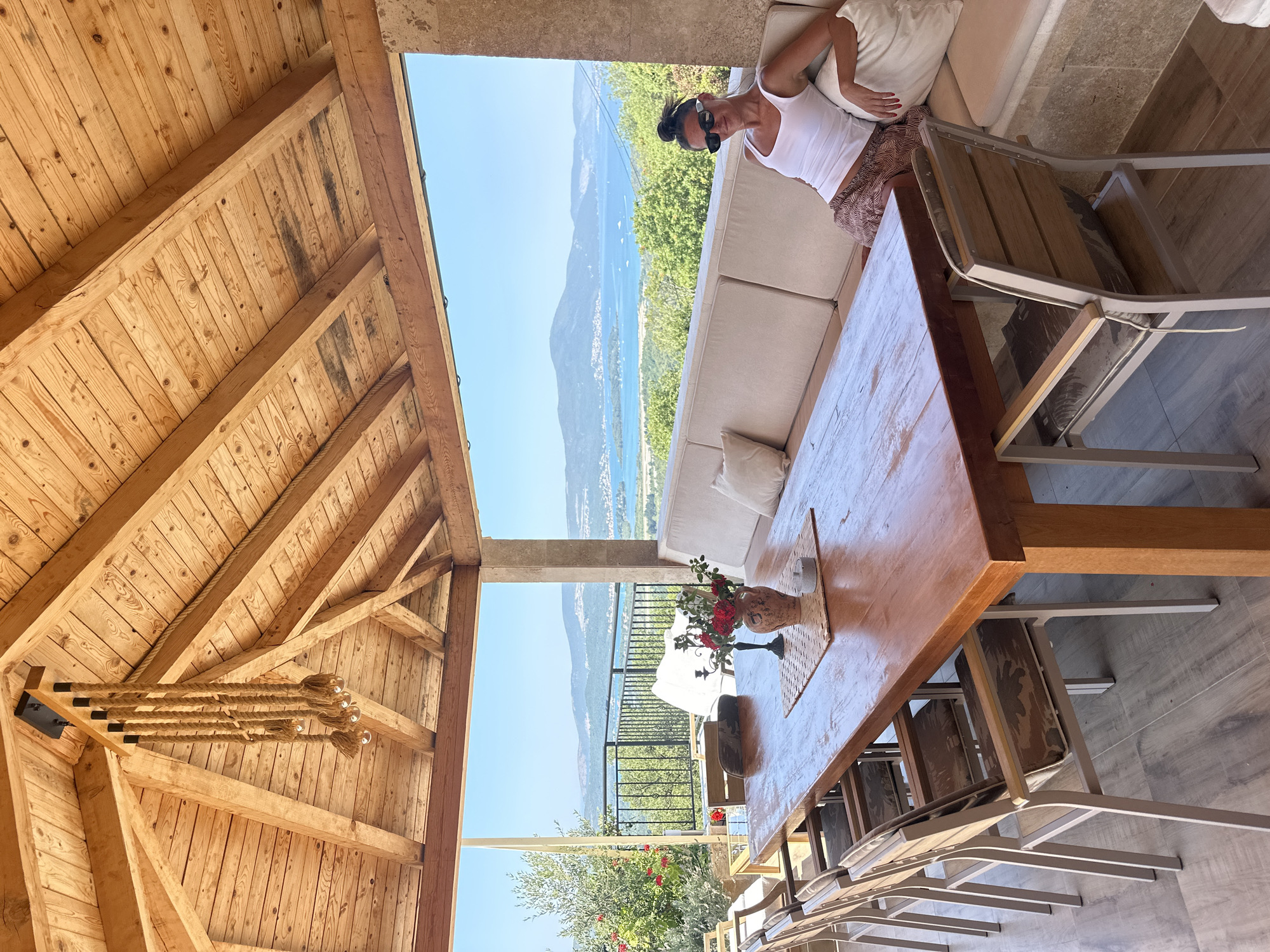In Kavač, Montenegro, an outdoor covered patio features a large wooden dining table with eight chairs. A woman wearing a white tank top and patterned skirt is seated on a built-in beige sofa, leaning against a pillow. The patio overlooks a scenic bay with islands and mountains under a clear blue sky. A chandelier made of rope and bulbs hangs from the wooden ceiling.