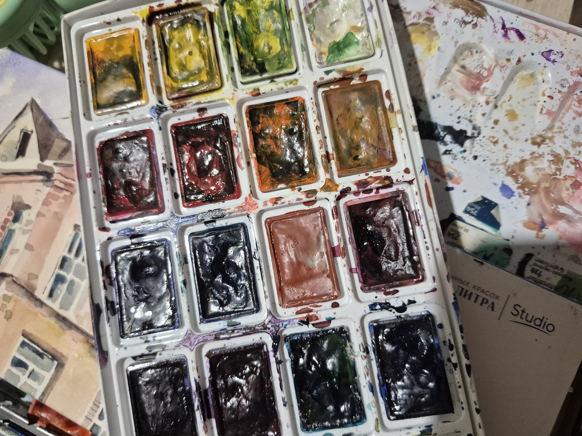The image shows a close-up, top-down view of a watercolor paint set with multiple pans of paint. The paint set is rectangular and appears to be made of white plastic. Some of the paint pans are full, while others are partially used, revealing swirls and textures. The colors visible include various shades of black, deep purple, red, orange, yellow, green, and a reddish-brown. Splatters and streaks of paint are visible on the white surface of the paint set, indicating recent use. In the background, out of focus, a portion of a watercolor painting depicting buildings can be seen, along with a white cardboard box with Cyrillic lettering that reads "Studio" and "ЛИПРА КРАСОК". The overall scene suggests an art studio or workspace. There are no people or specific locations within the city of Popovo, Bulgaria, identifiable in this image.
