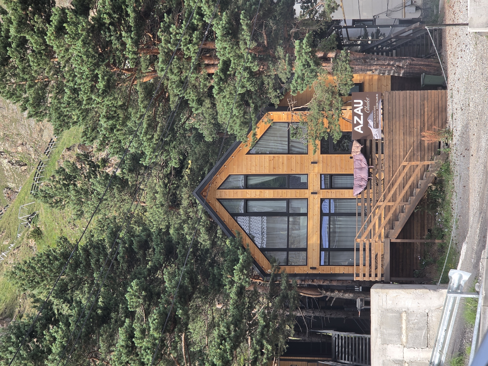 The image shows a modern wooden chalet named "AZAU Chalet" nestled amongst tall pine trees in the mountainous region of Terskol, Russia. The chalet has large windows, a sloping roof, and a wooden deck with stairs. A sign with the chalet's name and contact information is visible on the deck. In the background, a steep, grassy slope with a fence and what appears to be part of a ski lift or zipline can be seen. The foreground shows a gravel road and a concrete barrier. No people are visible in the scene. The overall environment suggests a natural, outdoor setting, likely a resort area in a mountainous landscape.