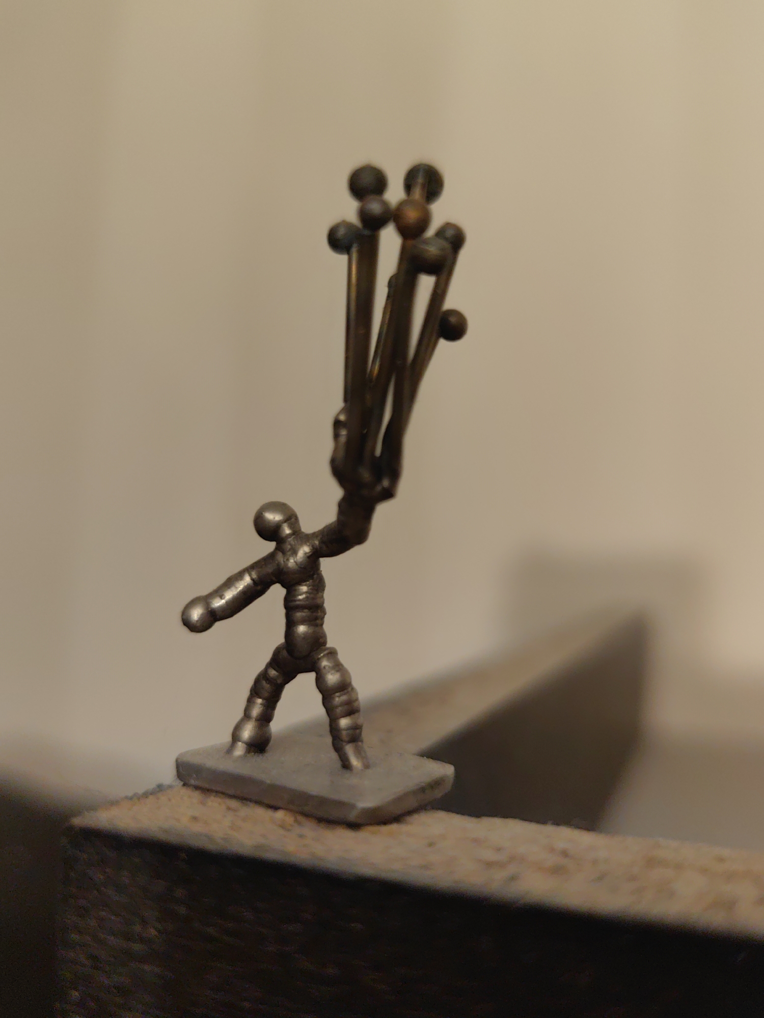 The image displays a close-up of a small, metallic figurine captured indoors in Sofia, Bulgaria. The central subject is a stylized human-like figure, rendered in a dull silver-grey metal. Its form is somewhat segmented, with rounded joints and a squat, sturdy posture. The figurine stands on a flat, rectangular base of the same metallic material.
With both arms raised, the figurine is holding aloft a bundle of approximately six thin, metallic rods. These rods are a darker, coppery-bronze color and each culminates in a small, spherical top. The pose suggests an action of carrying, presenting, or even wielding the bundle.
The figurine and its base are positioned near the edge of a textured, dark brown surface, possibly a piece of furniture or a shelf. The background is a soft, out-of-focus beige wall, providing a plain canvas that highlights the metallic sculpture. A faint shadow is cast on the wall to the right, suggesting an artificial light source from the left. The overall impression is that of a meticulously crafted miniature sculpture. There is no visible text in the image.