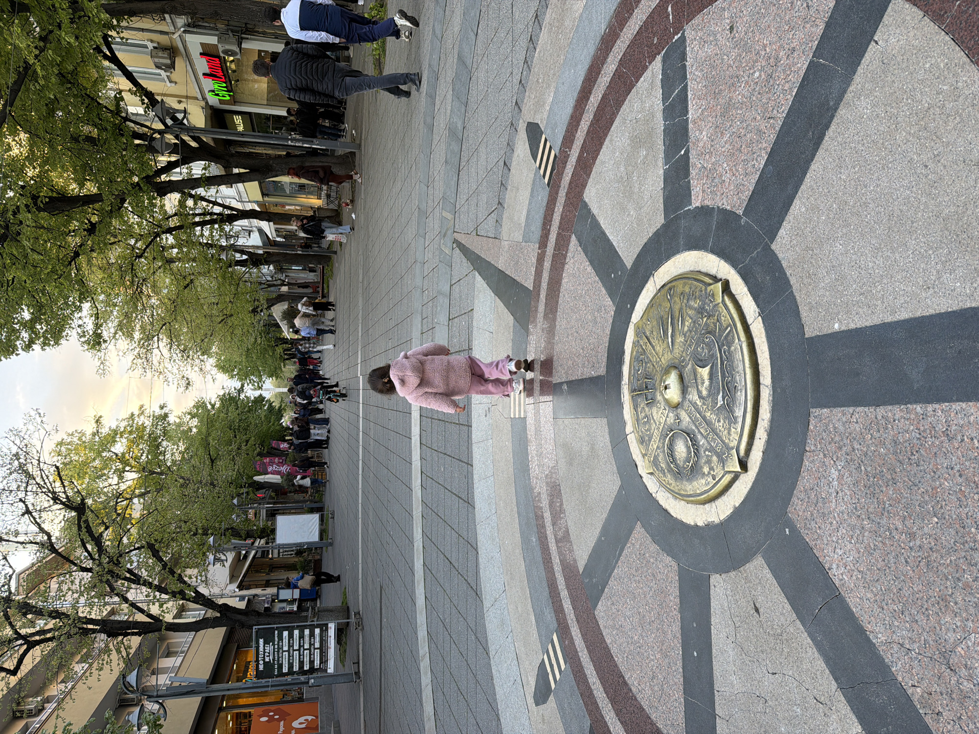 The image shows a street scene in what appears to be an urban area. A young girl wearing a pink outfit is standing on a large circular mosaic or design embedded in the pavement. The design features a central, possibly bronze or brass, medallion with inscriptions and patterns, surrounded by radiating lines of different colored stones.
Behind the girl, the street stretches out lined with trees, shops, and pedestrians. There is a sign mentioning "НЕФТОХИМИК" and other Bulgarian language text. Other signs visible are "GyroLand" and "PAUSE". There are trees overhead, partially obscuring the sky. The lighting suggests it is either early morning or late afternoon.