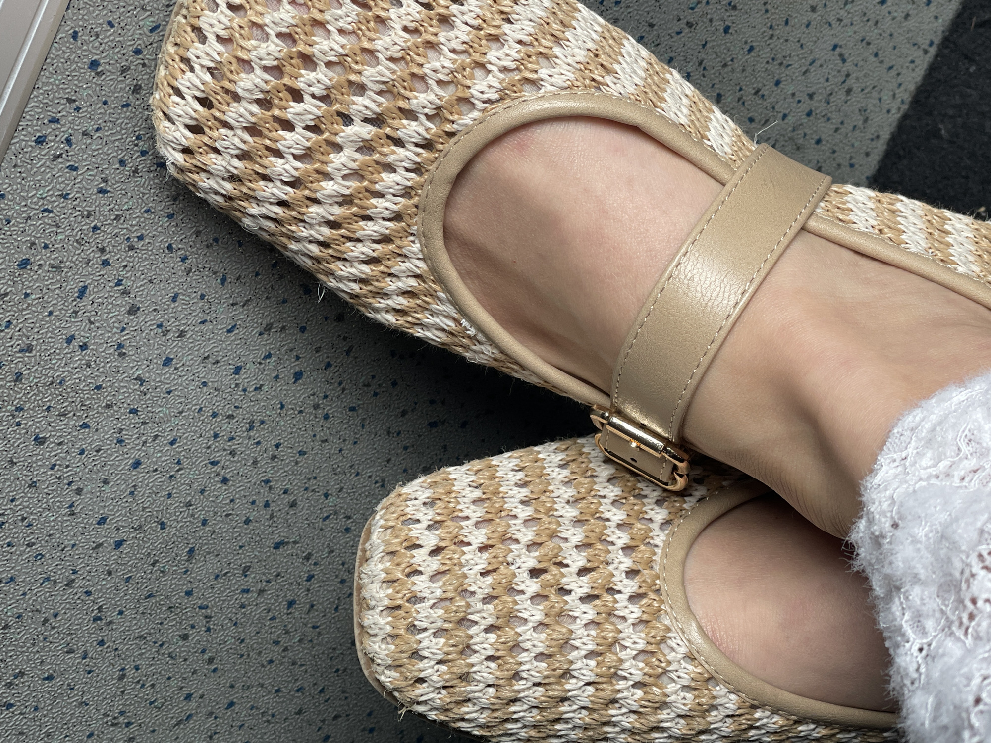 The media file shows a close-up, top-down view of two feet wearing tan and white woven flat shoes with a strap across the instep and a gold buckle. The shoes are placed on a textured gray surface with small blue and black speckles, possibly a floor or seating area. The right shoe is slightly closer to the viewer. A sliver of white lace trim is visible on the right side of the frame, near the ankle of the right foot. The image appears to be taken indoors, with neutral lighting. There are no people or text visible in the image. The context provided indicates the location is Ulmen, Germany, but this cannot be determined from the image itself.