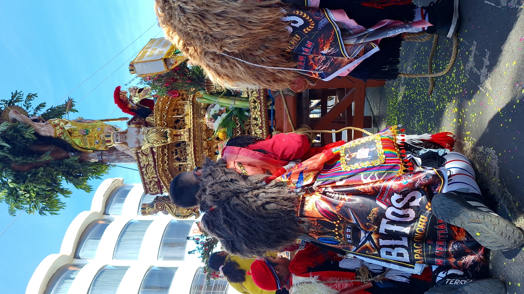The image depicts a religious procession likely in Wanchaq, Peru, with participants dressed in elaborate, traditional costumes. In the foreground, a person kneels, their back to the camera, wearing a large, shaggy, dark brown fur covering over their shoulders and back. They are adorned with a richly patterned skirt featuring colorful geometric designs and text, including "BLITOS" and "APACCONCHU". A framed image of a religious figure is attached to their clothing. Beside them, another individual is partially visible, wearing a similar furry costume and a scarf with text that includes "WANCHAQ".
In the background, a large, ornate float is prominent, featuring a sculpture of a figure resembling Jesus Christ standing on a cross, surrounded by a tree with green foliage. The float is elaborately decorated with gold accents and depicts a semi-nude female figure below the main sculpture. The procession is taking place outdoors on a paved surface, with small pieces of confetti scattered on the ground. In the far background, a modern building with curved white balconies is visible. The sky is clear and blue.