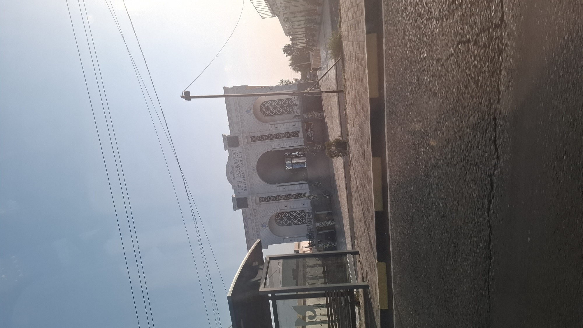 An outdoor, medium shot captures a building with ornate architecture and Arabic inscriptions, identified as "LIFE GARDEN SALTANAT SARAYI" in Shymkent, Kazakhstan. The building has a large central archway, decorative window grilles, and a pale, textured facade. To the left, a bus stop shelter with glass panels and benches is visible, partially obscuring the building's left side. In the foreground, a cracked asphalt road with yellow and black curb markings runs horizontally. Overhead, power lines crisscross against a clear blue sky with a bright sunlit haze on the right. The scene appears to be a street view during daytime.