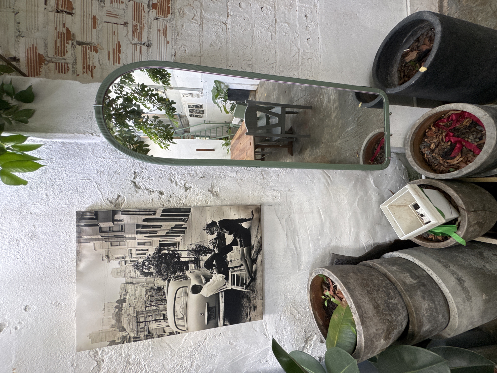 The image depicts an outdoor courtyard, likely in Phu Thuy Ward, Vietnam, with a distinctly eclectic and bohemian atmosphere. Dominating the left wall is a large black and white photograph of a city street scene, featuring San Francisco's iconic hilly terrain, vintage cars, and distinct architecture. The photograph appears to be a vintage print, perhaps representing a nostalgic aesthetic. In front of this photograph, a collection of weathered concrete planters are stacked, with one planter containing a young plant and another holding dried leaves and a red ribbon. To the right, a tall, oval-shaped green mirror is propped against a white textured wall, its surface reflecting a portion of the courtyard. The reflection reveals a wooden table with chairs, suggesting an outdoor dining or seating area. Beyond the mirror, a section of a red-bricked wall is visible, its texture contrasting with the smooth white wall. Green foliage hangs from the top of the frame, casting shadows and adding a natural element to the scene. The overall impression is one of a curated, lived-in space, blending art, nature, and repurposed objects.