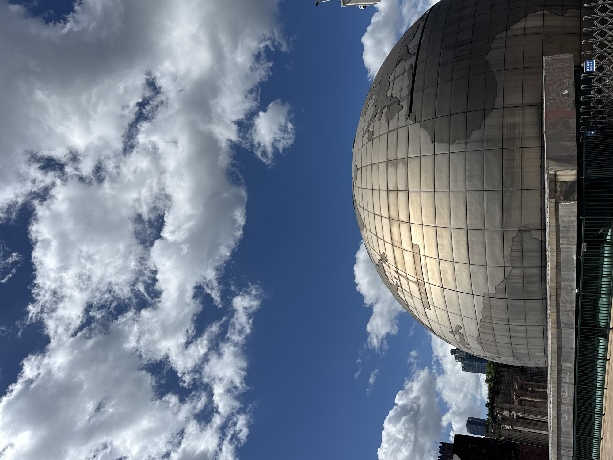 The image depicts a large, spherical structure resembling a globe, situated in Nanshan District, China. The globe is covered in metallic panels that form a map of the world. In the background, there are modern high-rise buildings and remnants of ancient architecture, including columns. The sky is a vibrant blue with scattered white clouds. A dark green metal fence runs along the foreground. There are no people visible in the image. The scene appears to be a public space or an outdoor exhibition.