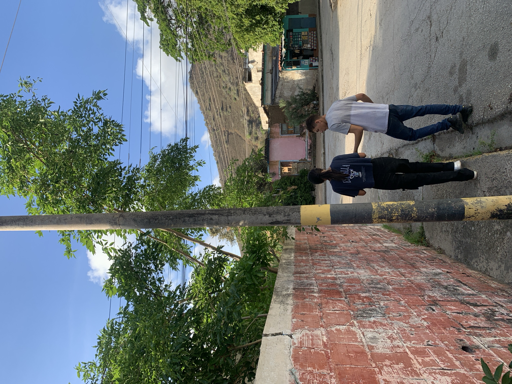 Two individuals, a woman with dark hair in a ponytail wearing a dark t-shirt and black pants, and a man with short dark hair wearing a grey t-shirt and blue jeans, are walking away from the camera on a paved street in Santa Ana, Venezuela. They are positioned to the left of a utility pole with black and yellow markings. To their left is a textured red brick wall. In the background, there are buildings, including a pink structure and a small storefront with a teal awning. Beyond the buildings, a steep, arid hillside rises under a blue sky with scattered clouds. Power lines are visible running across the sky and over the hillside. The overall scene depicts a street in a semi-arid, residential or commercial area.