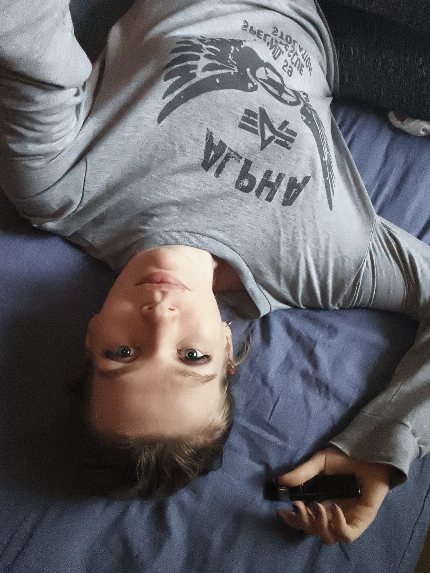 An indoor, eye-level shot captures a person lying on their back on a bed or couch, looking directly into the camera. The person's head is oriented towards the bottom of the frame, appearing upside down from the viewer's perspective. They have light skin, blue eyes, and light brown hair. Their expression is neutral and calm.
The person is wearing a long-sleeved, light grey t-shirt with a graphic design on the chest. The graphic includes a stylized logo resembling three horizontal bars, often associated with Alpha Industries. Adjacent to this logo and below it, large, mirrored, and upside-down text spells out "AH9JA". Above the logo, smaller, also mirrored and upside-down text is partially visible, reading "2ЬЕДЧОТЧОН 23" and to its right, partially "СПСИФС".
The person's right hand (viewer's left) holds a small, black electronic device, possibly a smartphone or vape, with purple-painted fingernails visible. The background consists of a dark blue fabric, likely a sheet or blanket, covering the surface they are lying on. The lighting suggests an indoor setting, but the time of day is not discernible. The overall scene conveys a casual, relaxed moment in Русский Вожой, Russia.