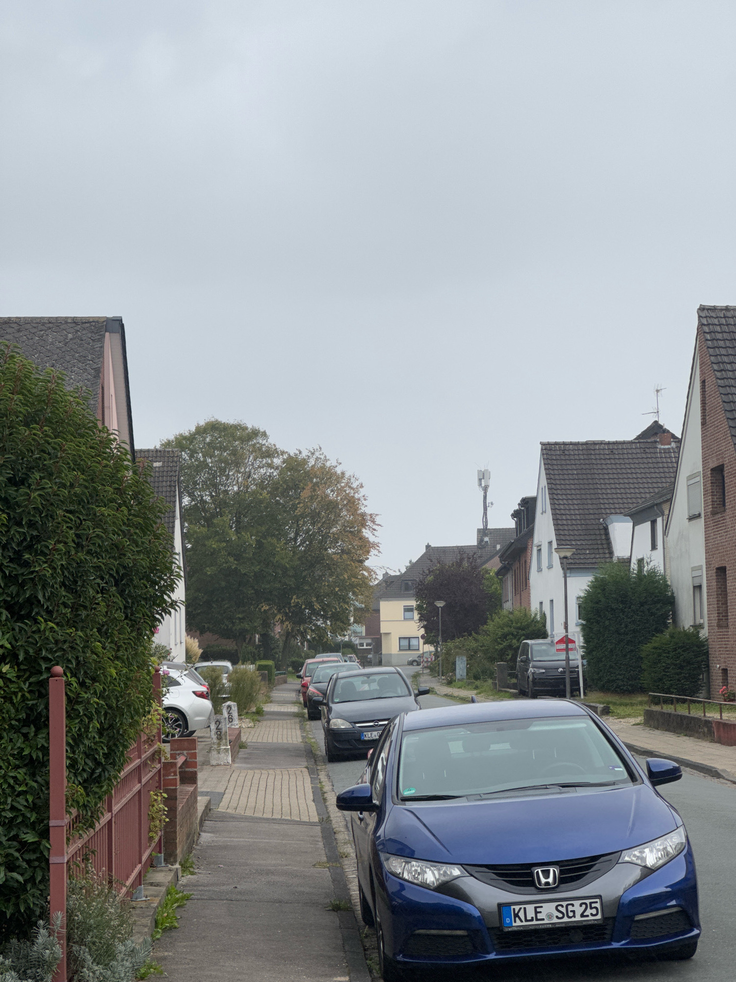 The image depicts a residential street in Cleves, Germany, on an overcast day. The foreground is dominated by a blue Honda Civic parked on the street, with its license plate reading "KLE SG 25". To the left, a sidewalk is visible, bordered by a red metal fence and a dense green hedge. Further down the street, several other cars are parked, including a dark SUV and other dark-colored passenger vehicles. A white SUV is parked on the side of the road near a sidewalk. The street is lined with houses, characterized by pitched roofs and brick or white facades. Mature trees with green and some autumnal foliage are scattered throughout the scene. In the distance, a cell tower is visible. The overall impression is that of a quiet, suburban residential area.