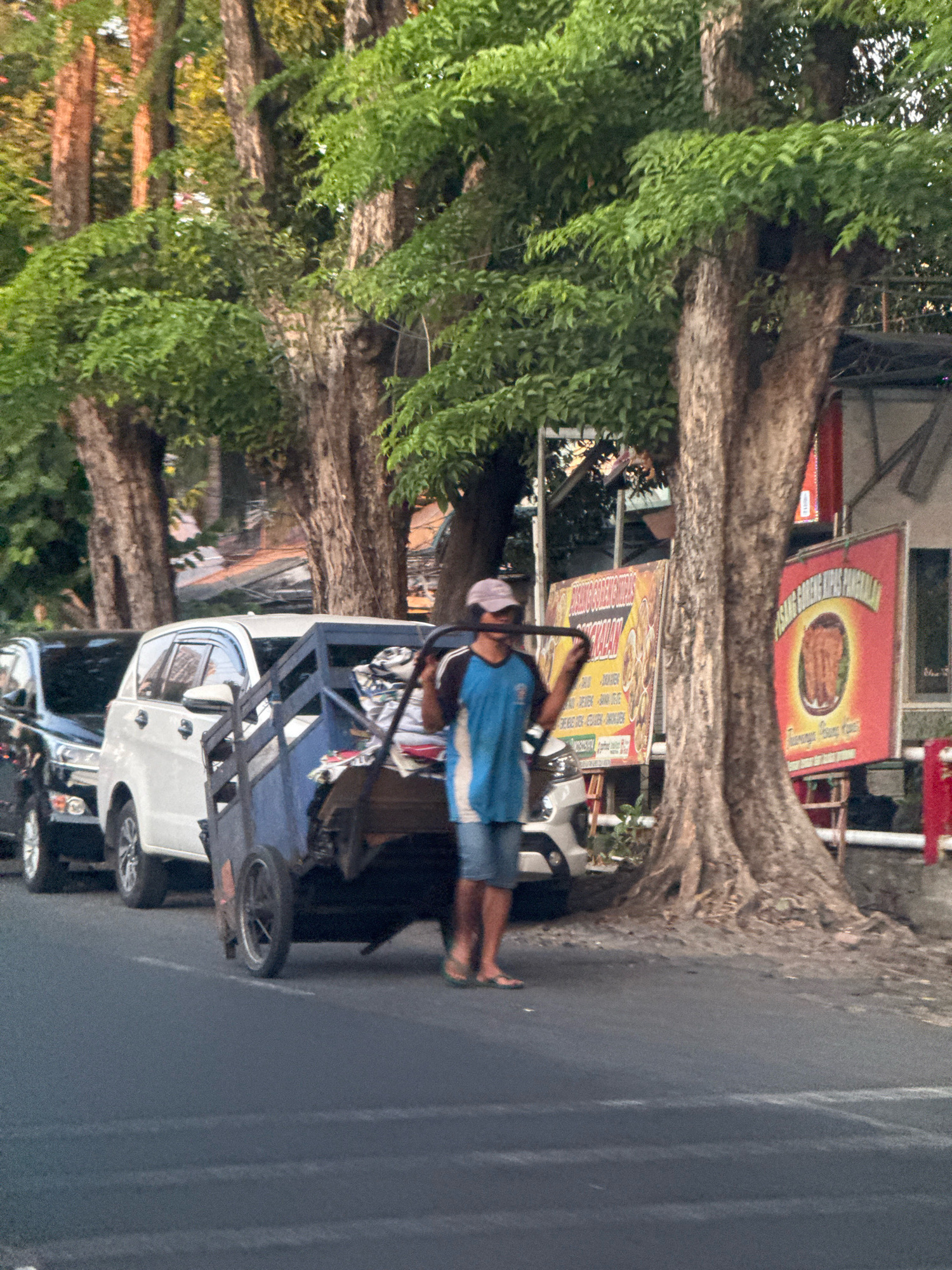 In Surabaya, Indonesia, a person is seen pushing a cart loaded with what appears to be fabric or laundry. The cart has two wheels and is being pulled by a yoke over the person's shoulders. The person is wearing a blue t-shirt, denim shorts, and sandals. They are also wearing a light-colored cap and a face mask. The scene is set on a street lined with large trees, and there are cars parked along the side of the road. A white SUV is visible in the foreground, as well as a black car. In the background, there are buildings with signs, suggesting a commercial area. The lighting indicates it is either early morning or late afternoon. The overall impression is a street scene in a bustling Indonesian city.